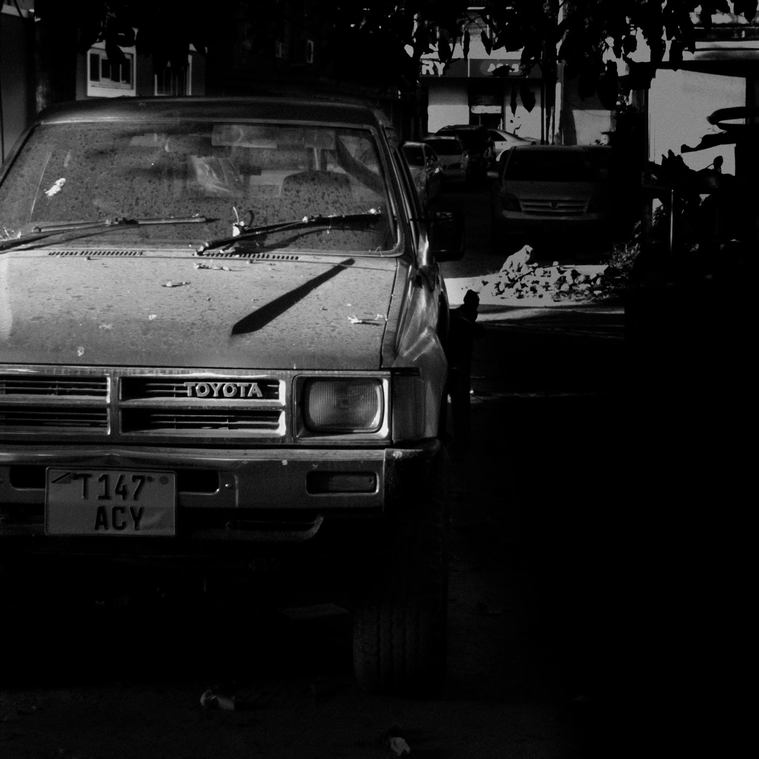 An old dusty Toyota pickup truck parked on a street with other cars in the background, with leaves on the windshield and decorations hanging above.