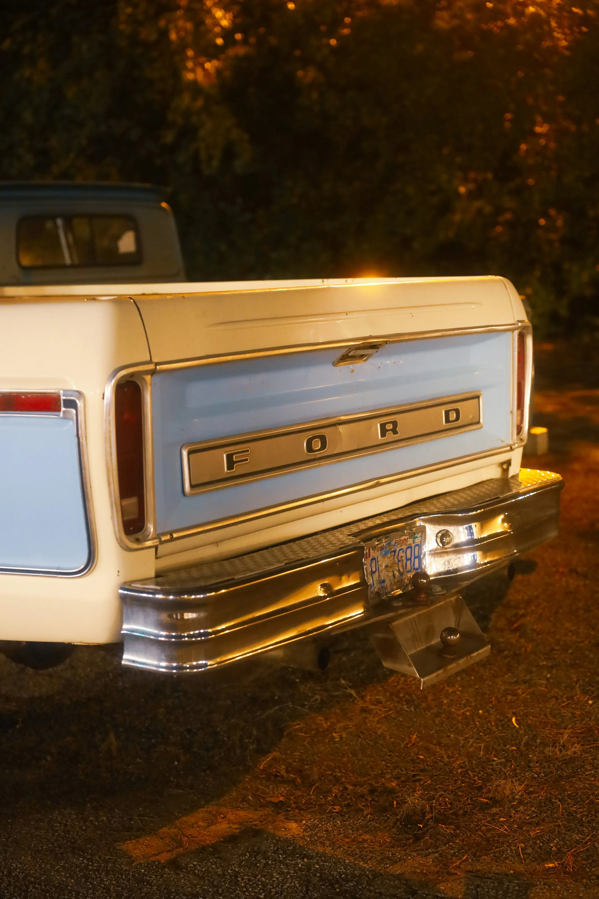 Close-up of the rear of a vintage Ford truck, showing the Ford emblem, rear bumper, and license plate, parked on a dark street at dusk.
