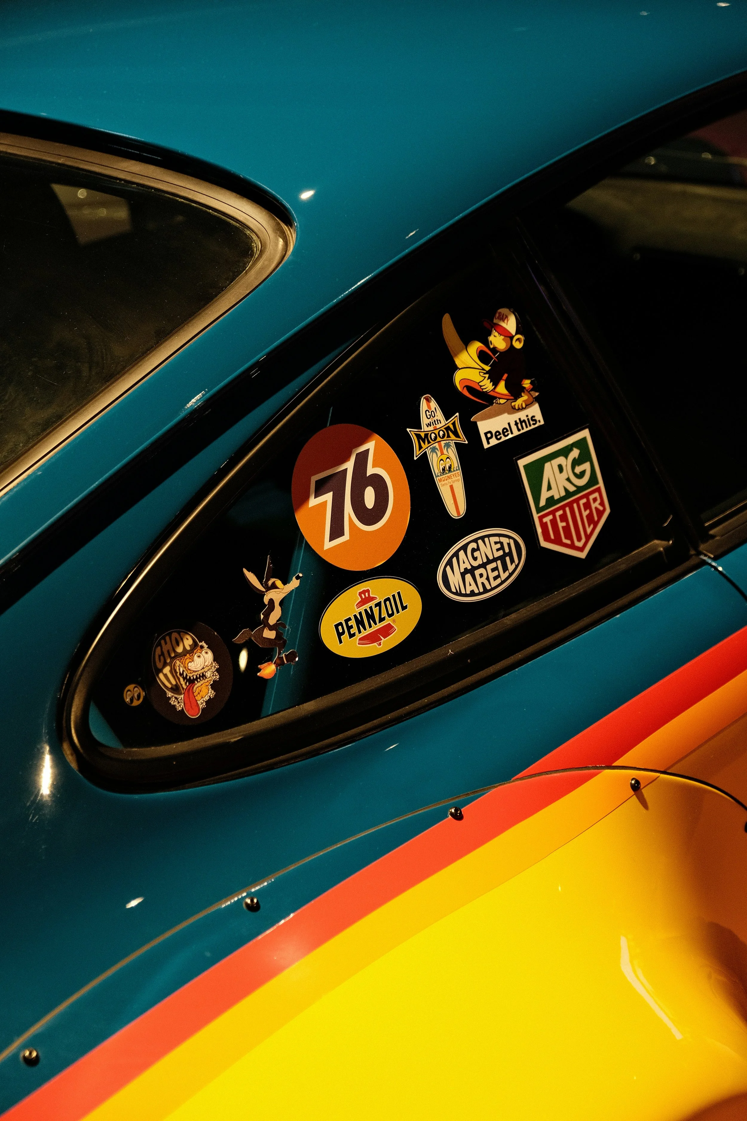 Close-up of a vintage car's rear window with various colorful stickers, including racing brands, cartoon characters, and playful messages.