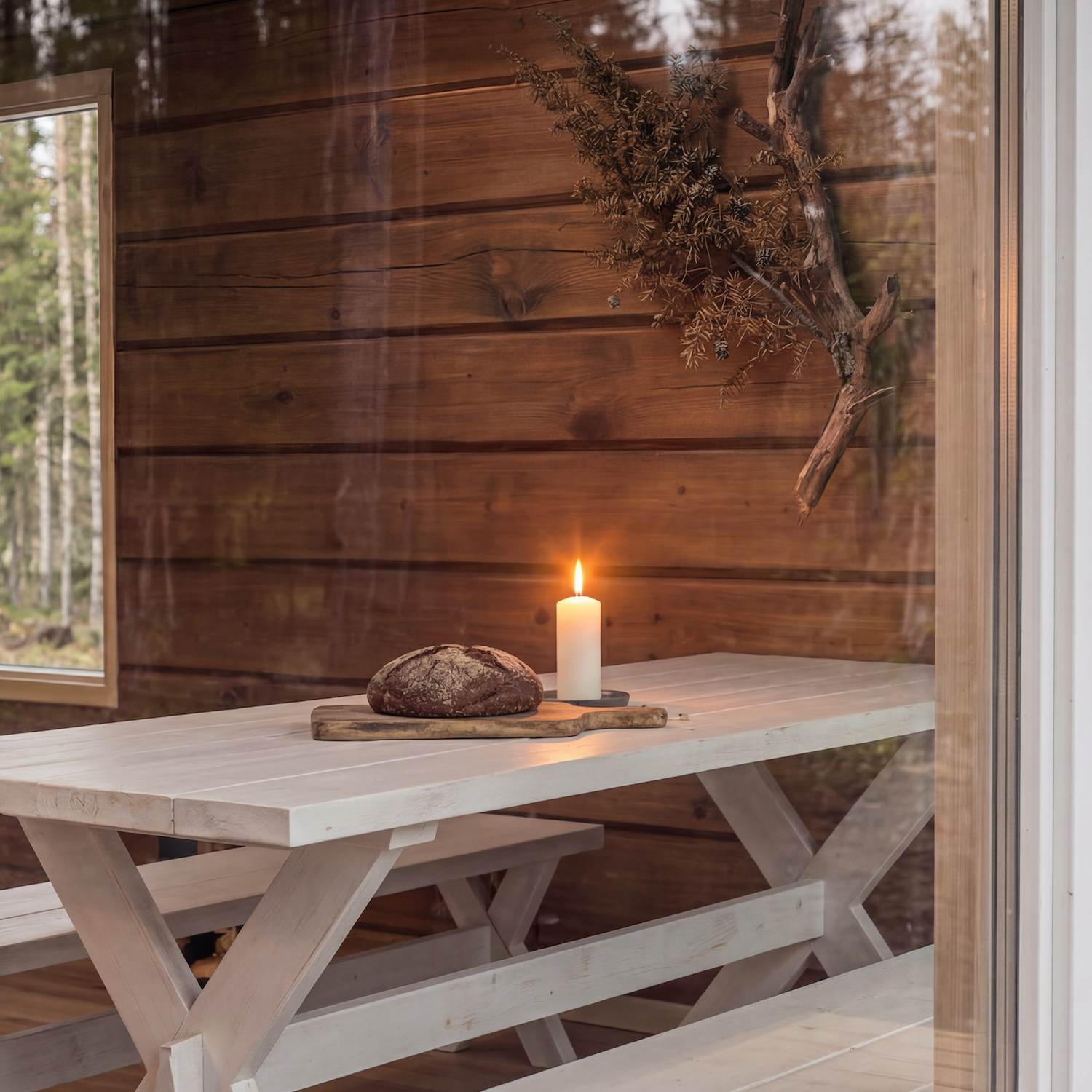 Tuolin päällä on leipä, kynttilä ja leikkuulauta, taustalla puinen seinä ja ikkunasta näkyy metsää.