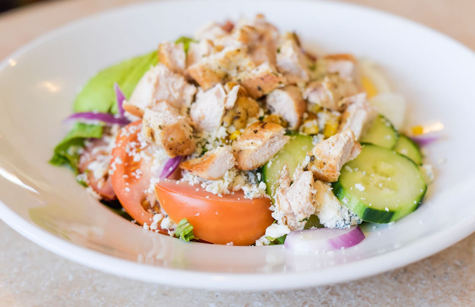 Close-up of a fresh chicken Caesar salad with tomatoes, cucumbers, red onions, croutons, grated cheese, and lettuce in a white bowl.