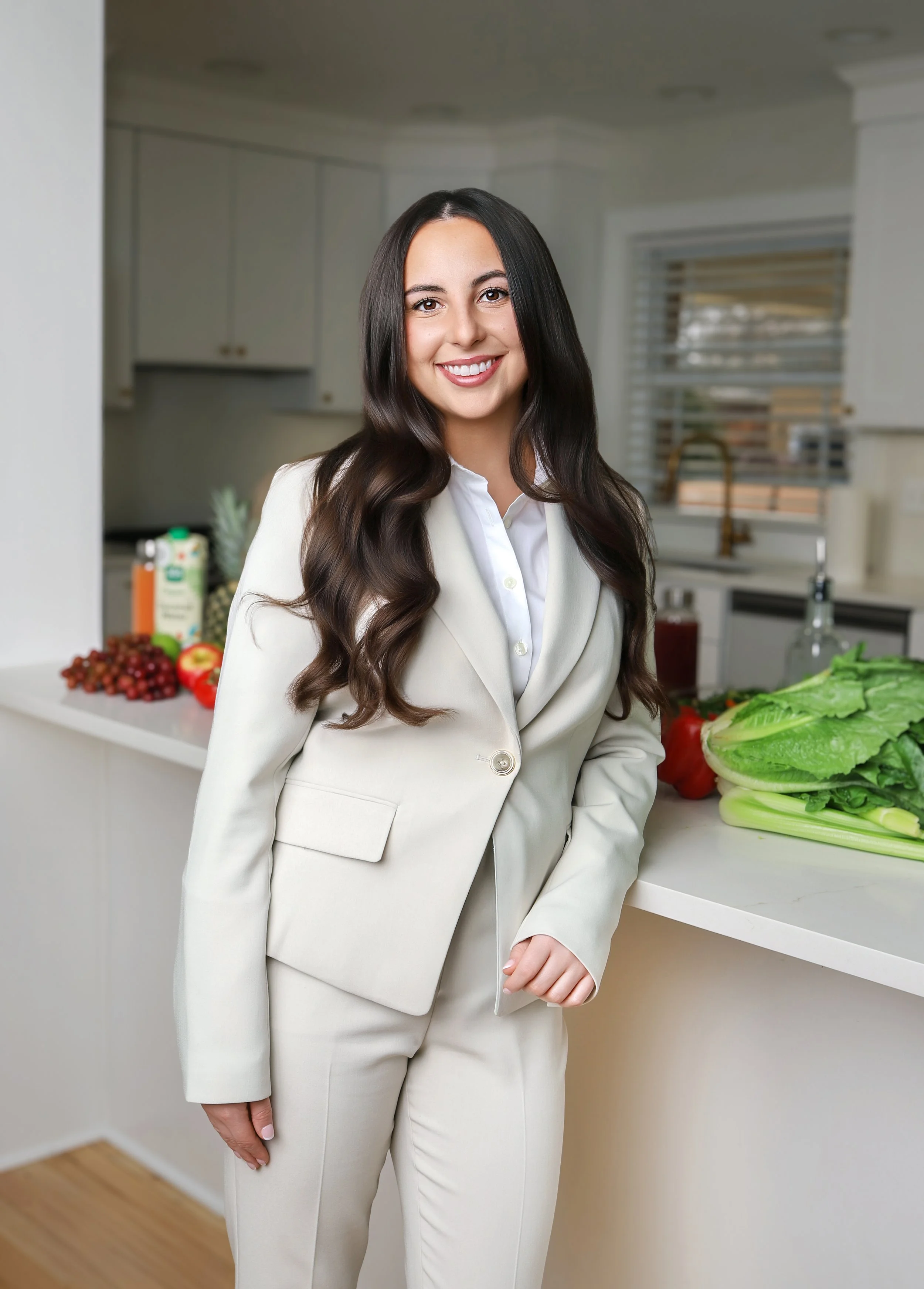 A woman with long, dark hair smiling in a kitchen, dressed in a light-colored suit with white shirt, standing next to a white countertop with vegetables and fruits in the background.