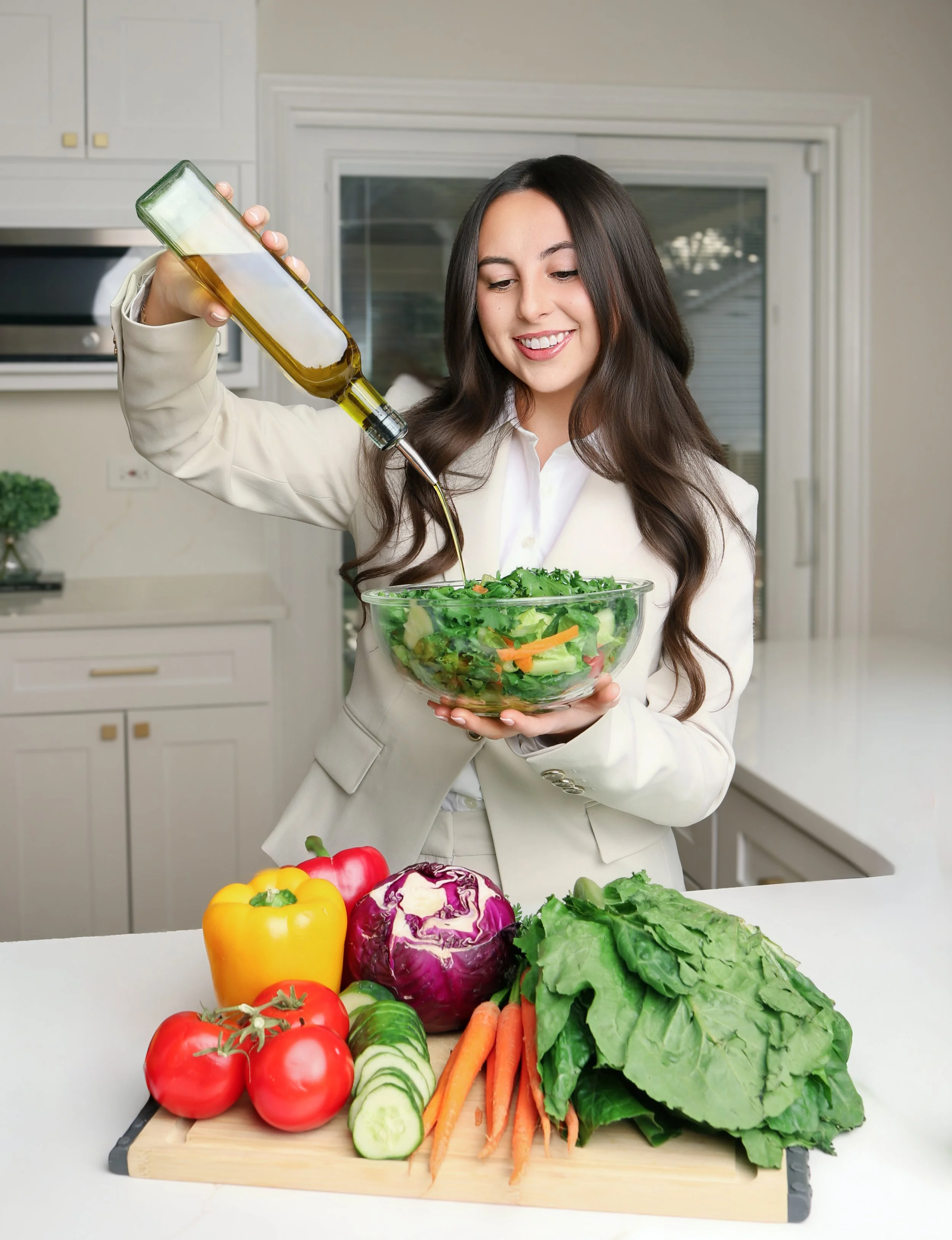 A woman in a white blazer pouring olive oil into a large bowl of salad, with fresh vegetables including tomatoes, peppers, lettuce, carrots, and cucumber on the kitchen counter.