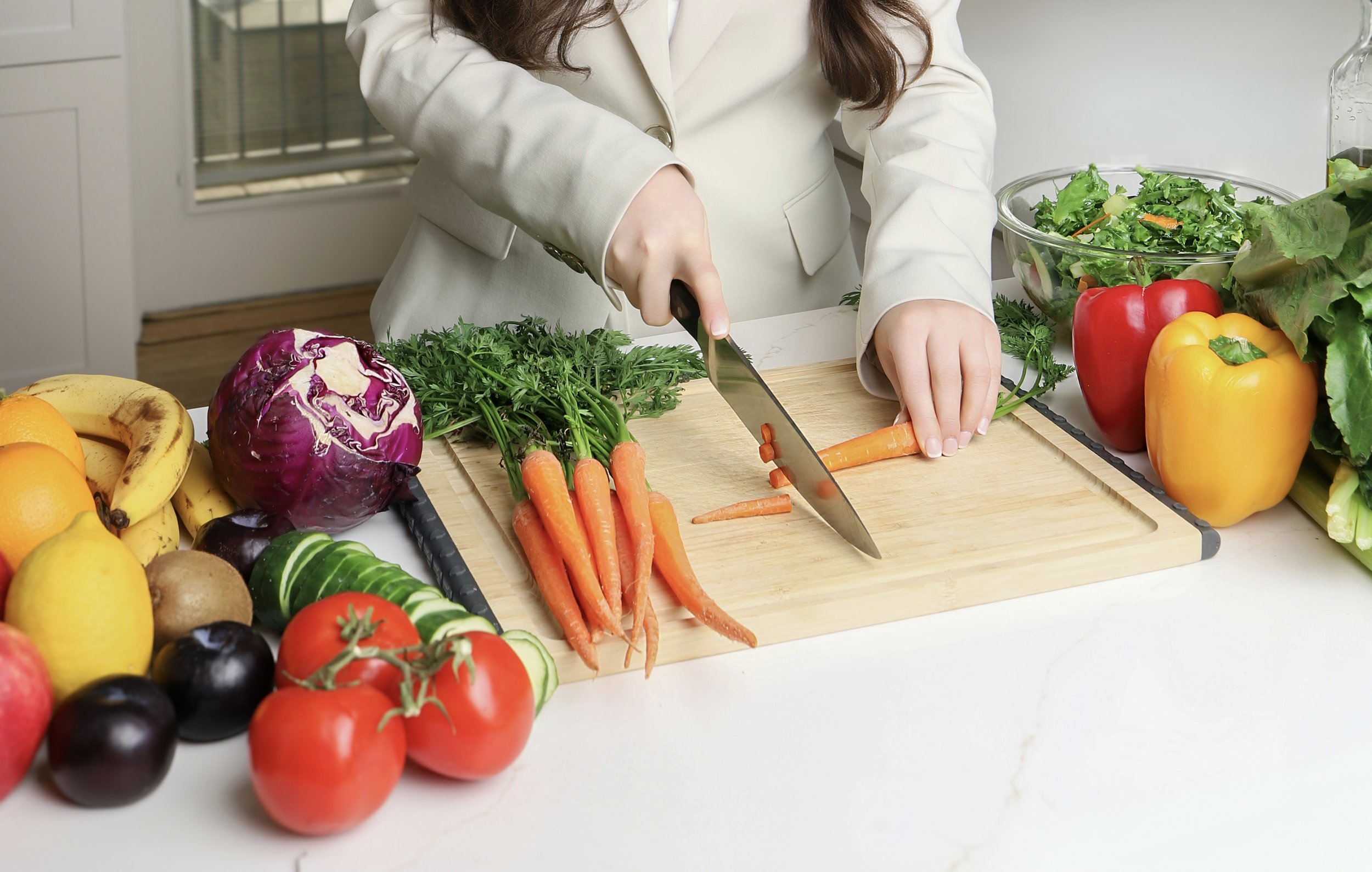 Person slicing carrots on a cutting board surrounded by various fresh vegetables and fruits, including lettuce, red and yellow bell peppers, tomatoes, and bananas in a kitchen.