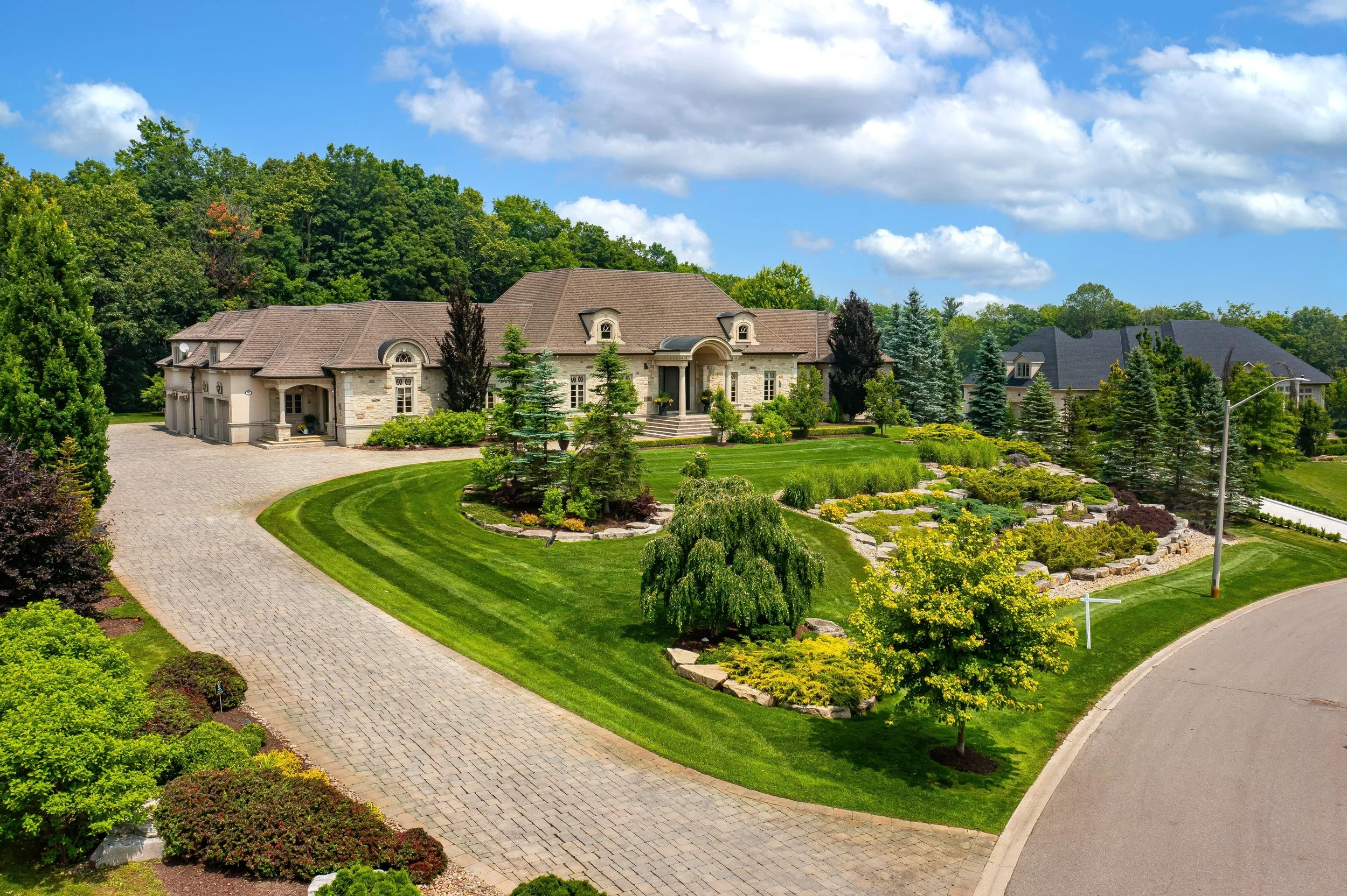 Large mansion with stone exterior, surrounded by well-maintained lawn, trees, and colorful bushes. Paved driveway curves around the property under a blue sky with white clouds.