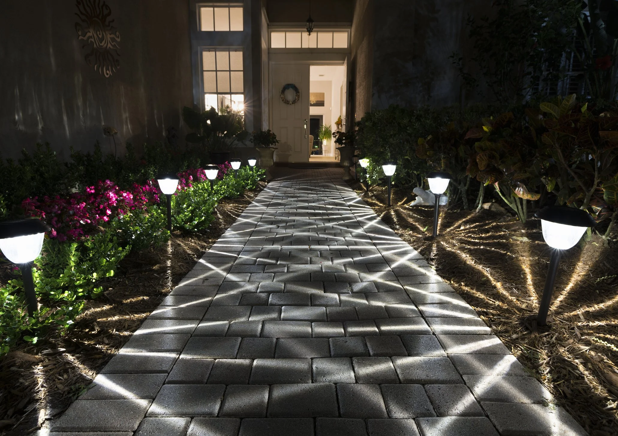 Nighttime view of a brick walkway illuminated by solar-powered pathway lights, leading to a house entrance decorated with a wreath, with flowerbeds on both sides containing pink flowers and green foliage.