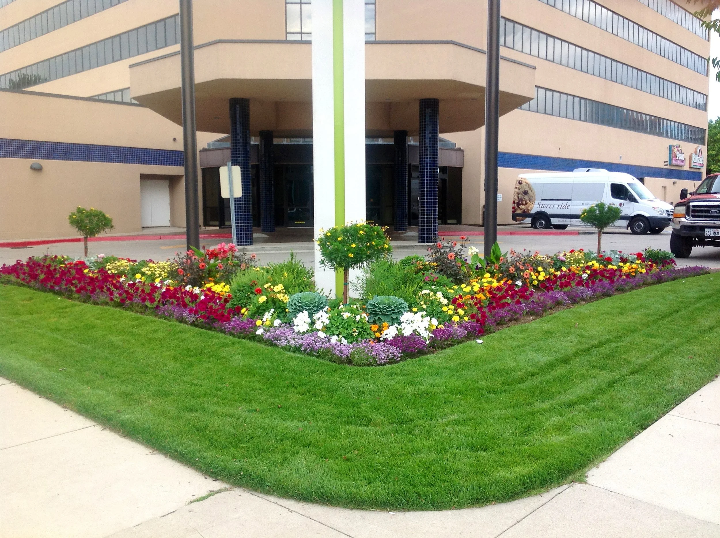 Colorful flowerbed with various flowers and plants in front of a beige building with blue accents, parking lot, and parked vehicles including a white van and a pickup truck.