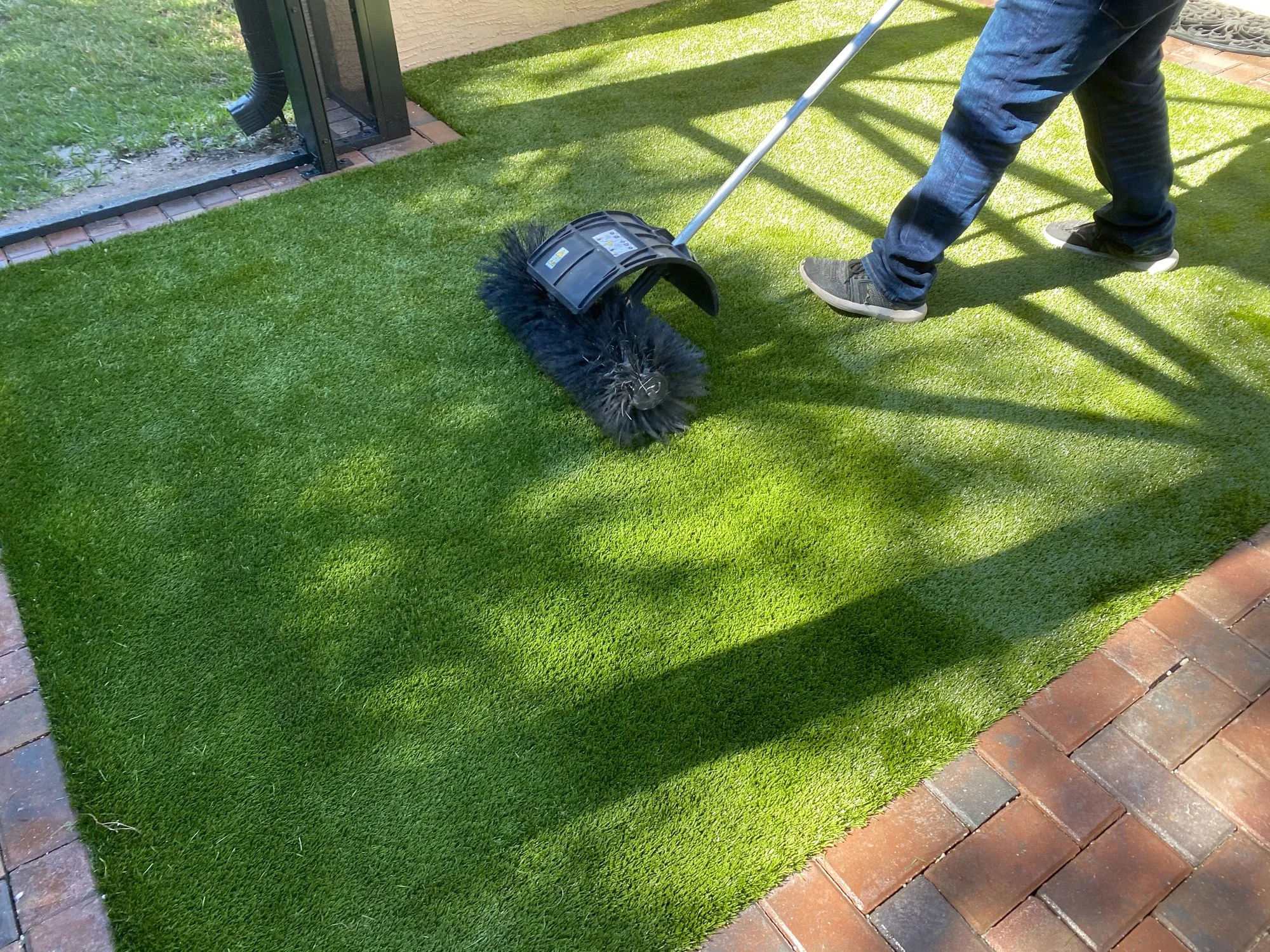 Person using a large broom to sweep artificial grass on a patio, with sunlight and shadows.