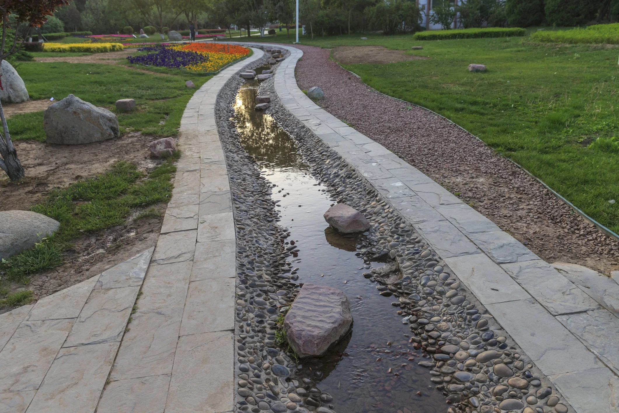 A winding stone pathway with a narrow, pebble-lined water stream running along the side, surrounded by green grass, rocks, and colorful flower beds in a park setting.