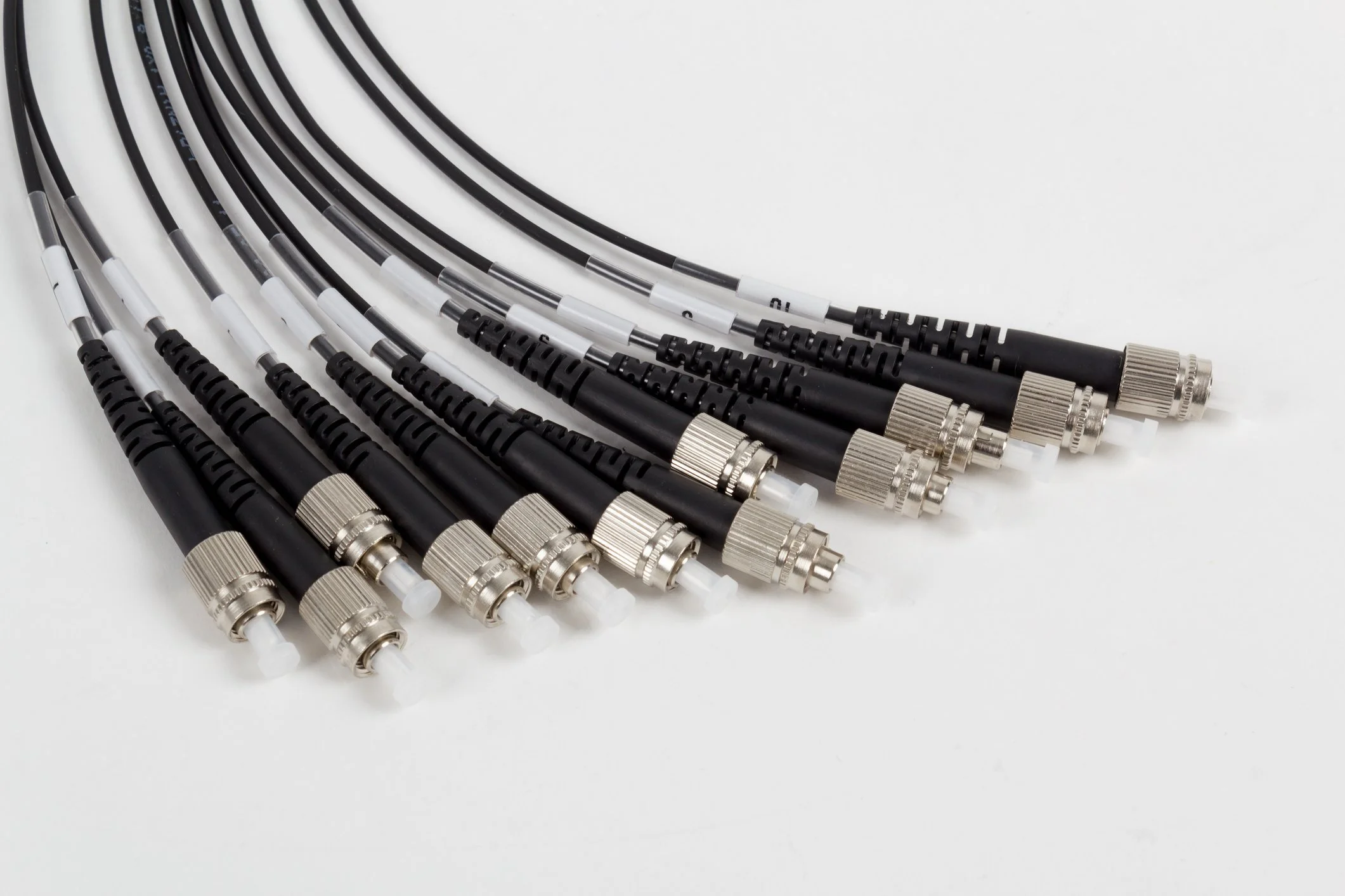 Multiple fiber optic cables with black and silver connectors arranged in a fan shape on a white surface.