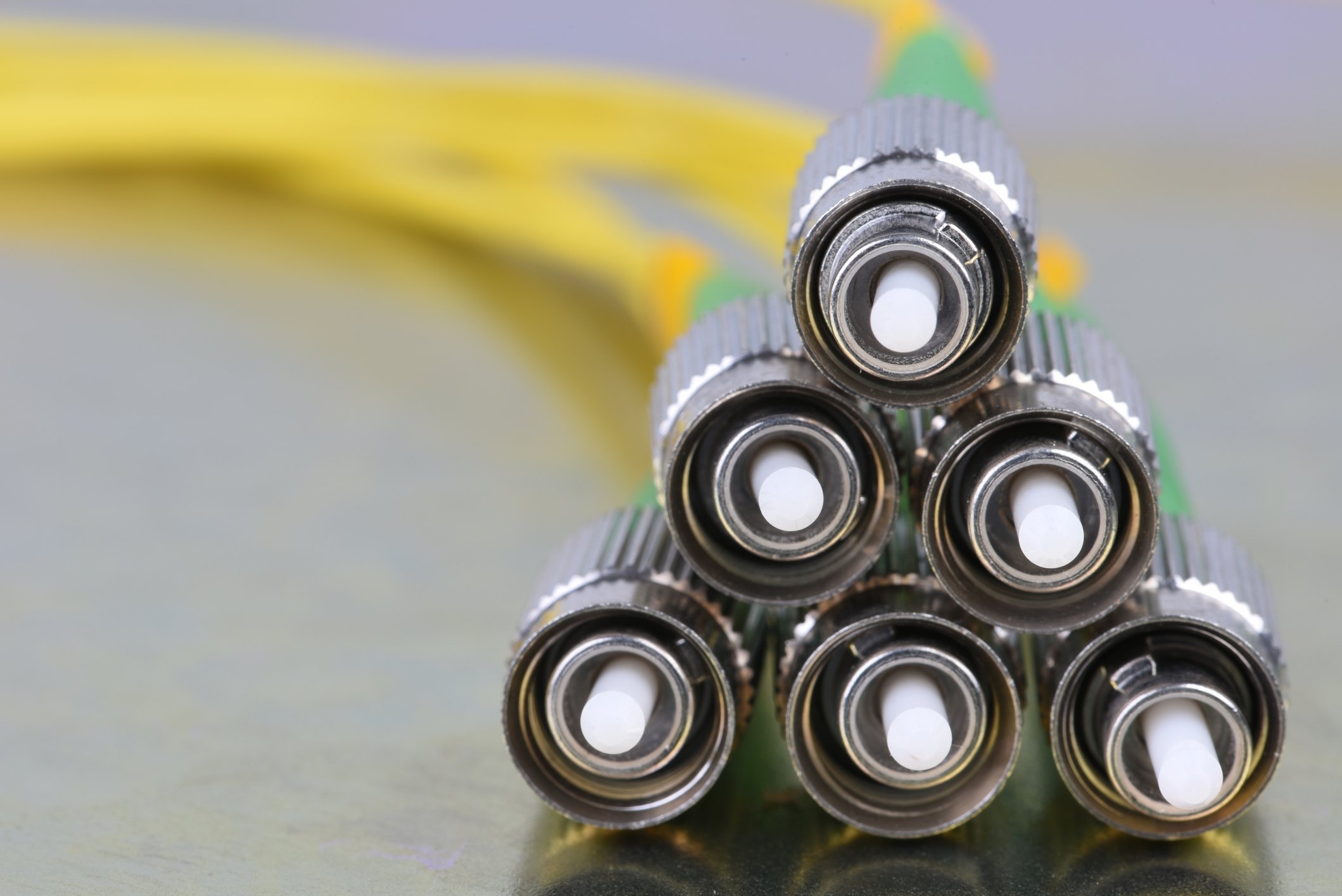 A close-up of seven fiber optic cables with metal connectors arranged in a pyramid shape on a flat surface, with yellow cables in the background.
