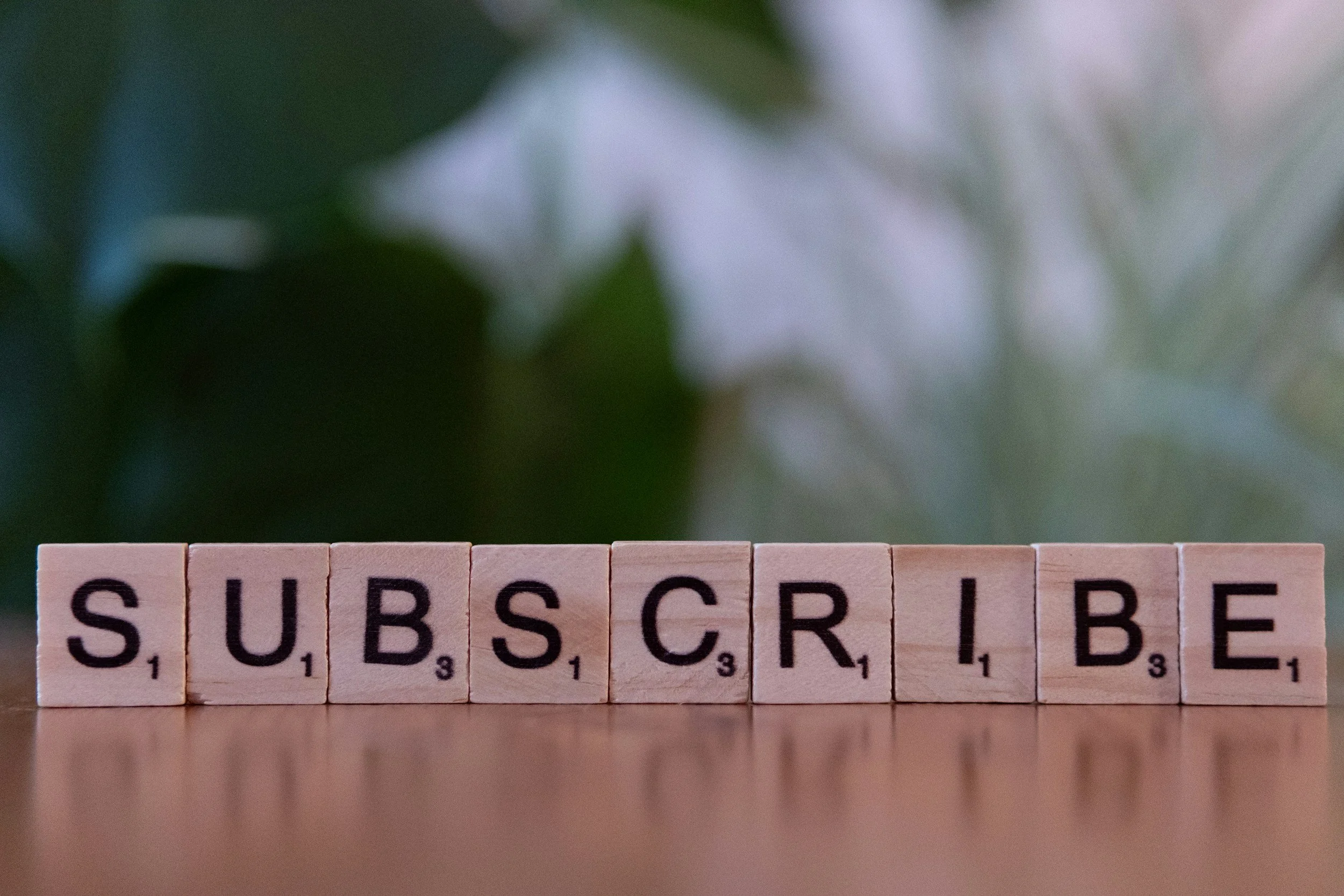 Scrabble tiles spelling 'SUBSCRIBE' on a wooden surface with a blurred green and white background.