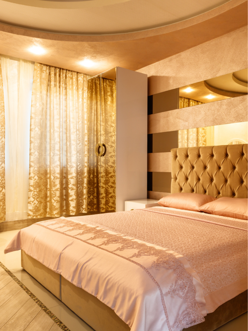 A luxurious bedroom with a large bed featuring a tan tufted headboard, peach bedding, and matching pillows. Gold patterned curtains cover a window, and the room has warm lighting reflecting off textured walls with horizontal striped design.