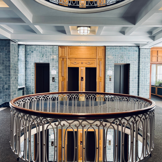 Interior of a building with two elevators framed by wooden doors, blue tiled walls, a decorative railing on the upper level, a ceiling light fixture, and a staircase opening with a railing in the foreground.