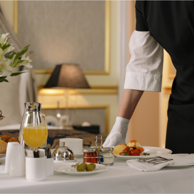 A waiter setting a breakfast or brunch table with orange juice, toast, eggs, and fruit in a hotel or restaurant dining area.