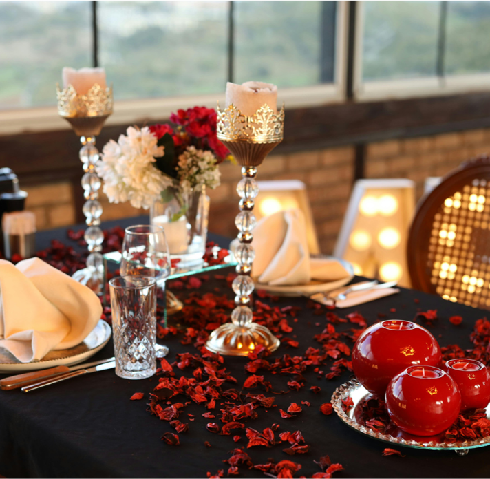 Elegant table decorated with red flower petals, white napkins, glassware, and candle holders, set for a special occasion.