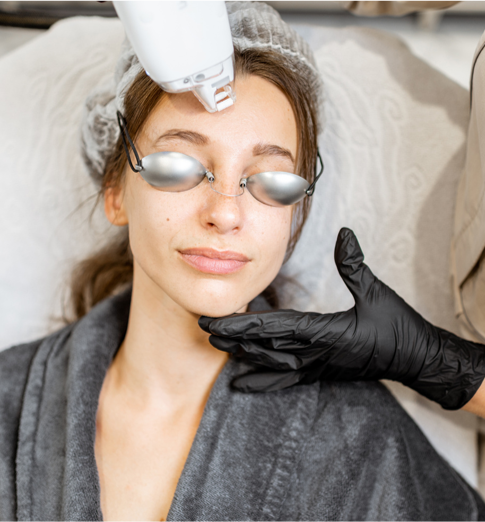 Woman receiving a cosmetic treatment with a device on her forehead, wearing protective eye goggles, and a technician with black gloves touching her chin.