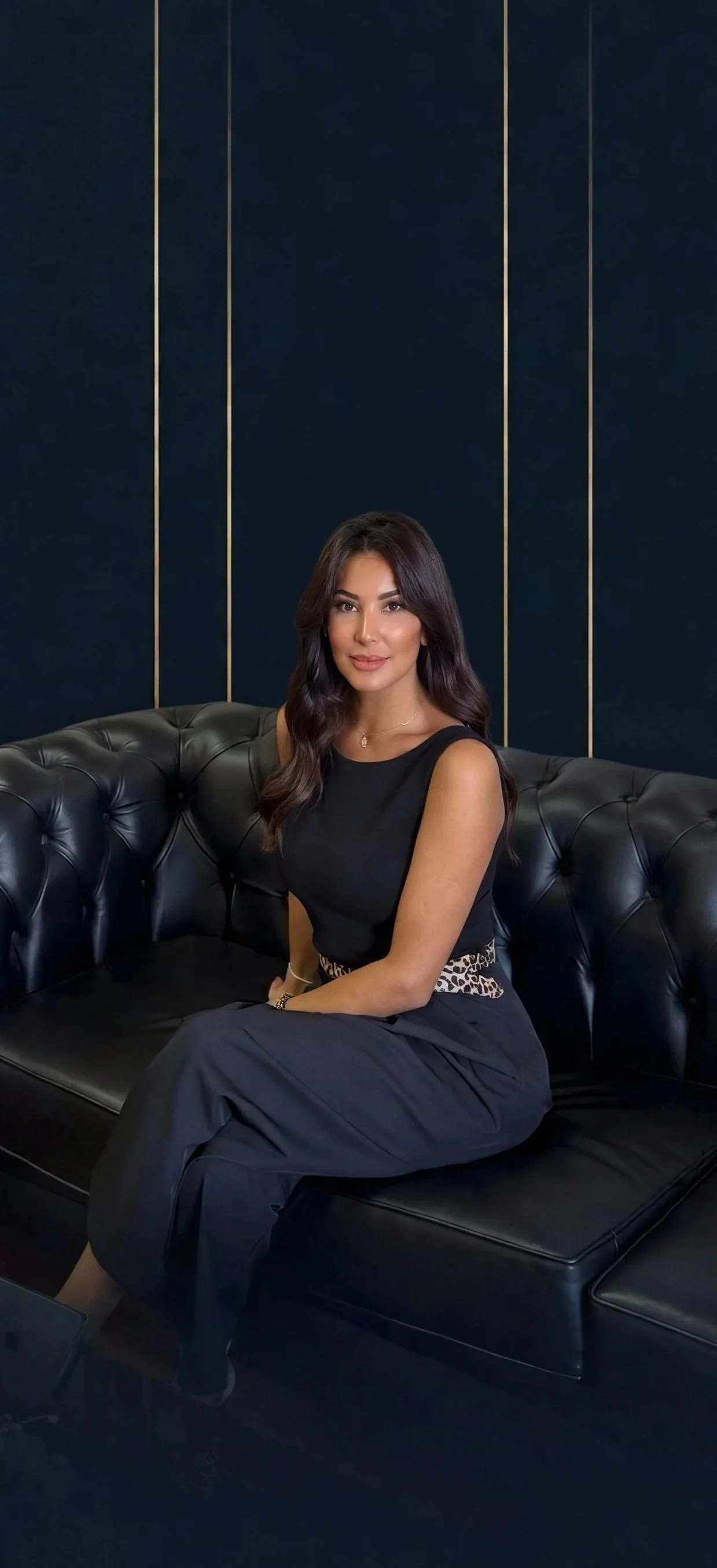 Founder and owner of the company '3f60' A woman with long dark hair, wearing a black sleeveless top and black pants, is sitting on a black leather tufted sofa in front of a dark blue wall with vertical gold stripes.