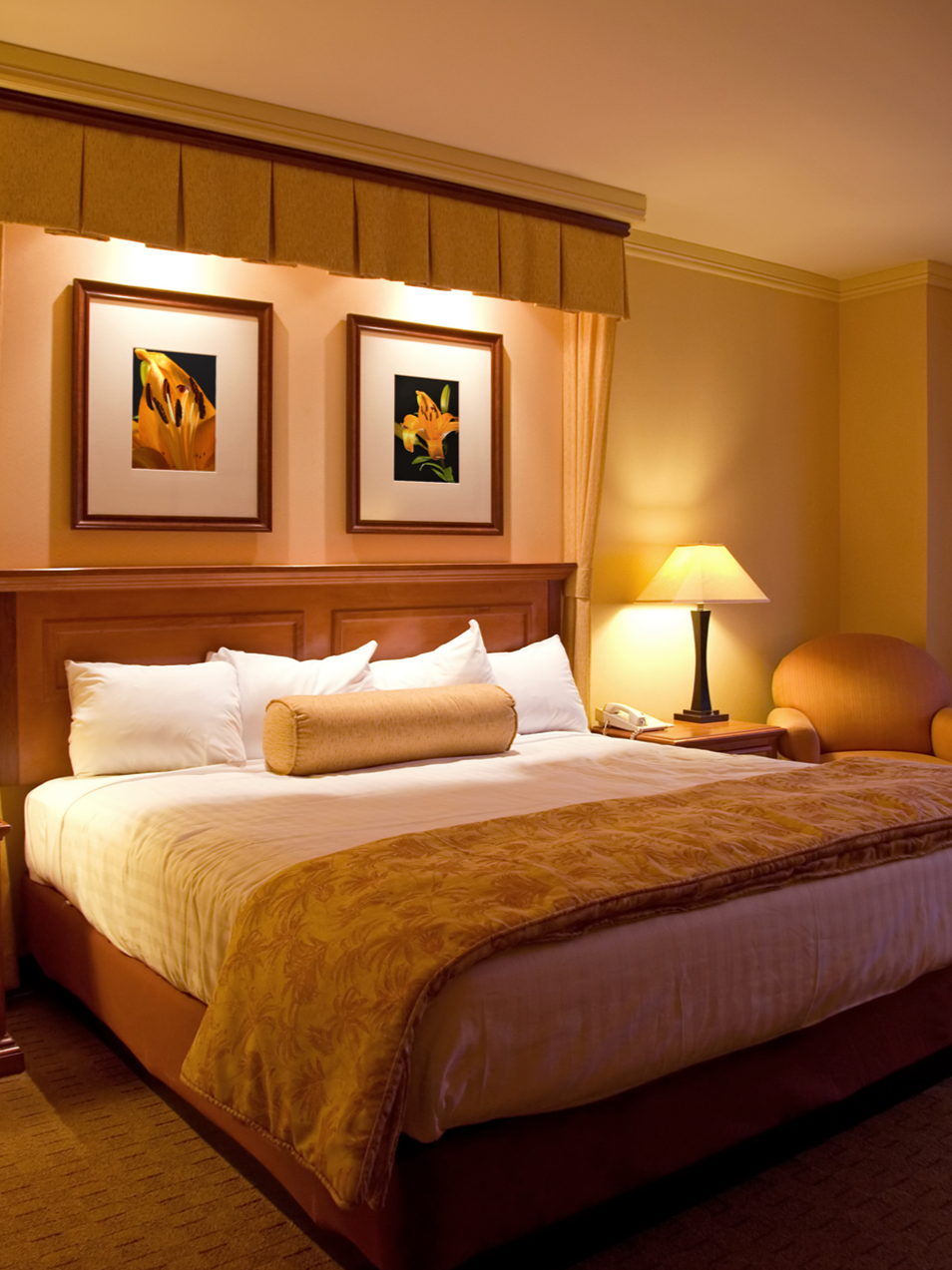 A hotel room with a large bed, white pillows, a cylindrical bolster pillow, and a decorative bedspread. There is a wooden headboard, framed flower pictures above the bed, a bedside lamp, a phone, and an armchair.