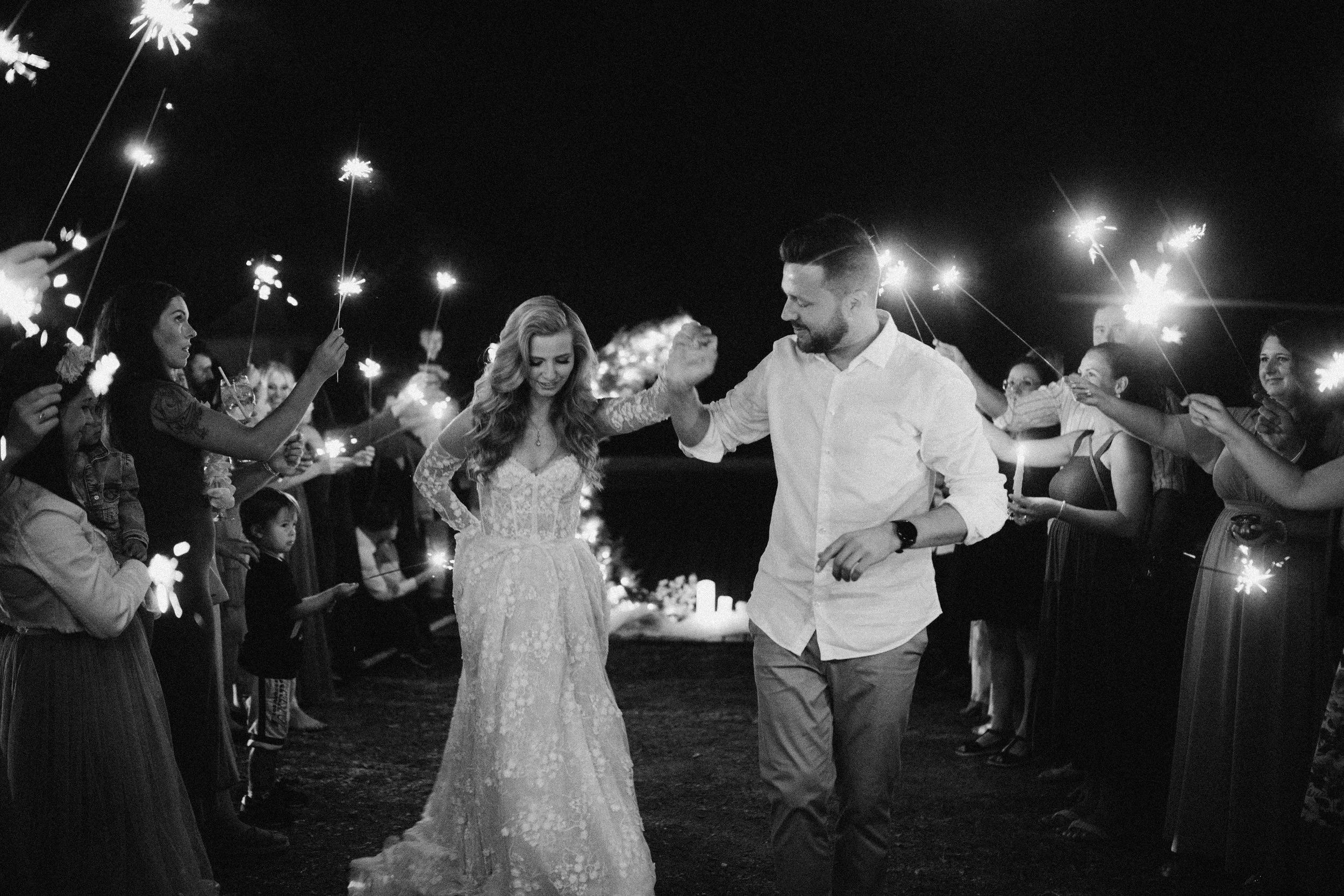 A bride and groom dancing at night surrounded by guests holding sparklers.