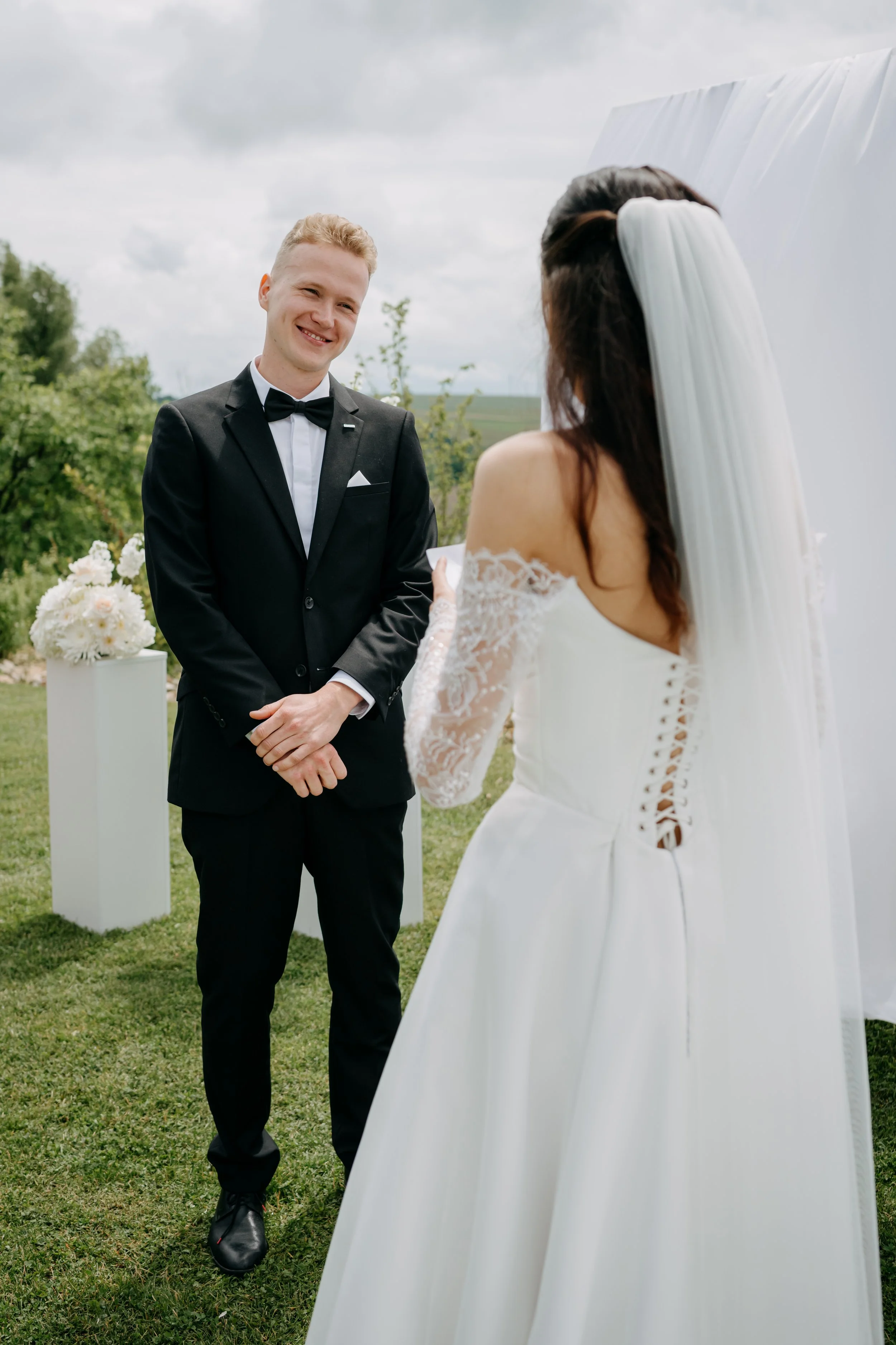 A groom in a black tuxedo and bow tie standing outdoors, smiling while looking at a bride dressed in a white wedding gown with lace sleeves and a long veil, during a wedding ceremony on a cloudy day with greenery in the background.