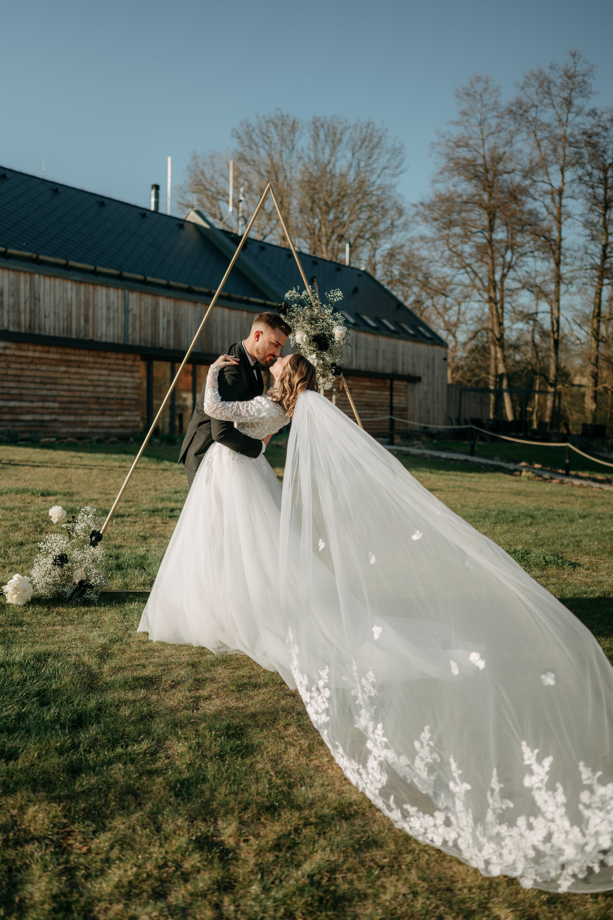 A bride and groom kiss outdoors under a triangular wedding arch decorated with flowers, with a rustic building and trees in the background.