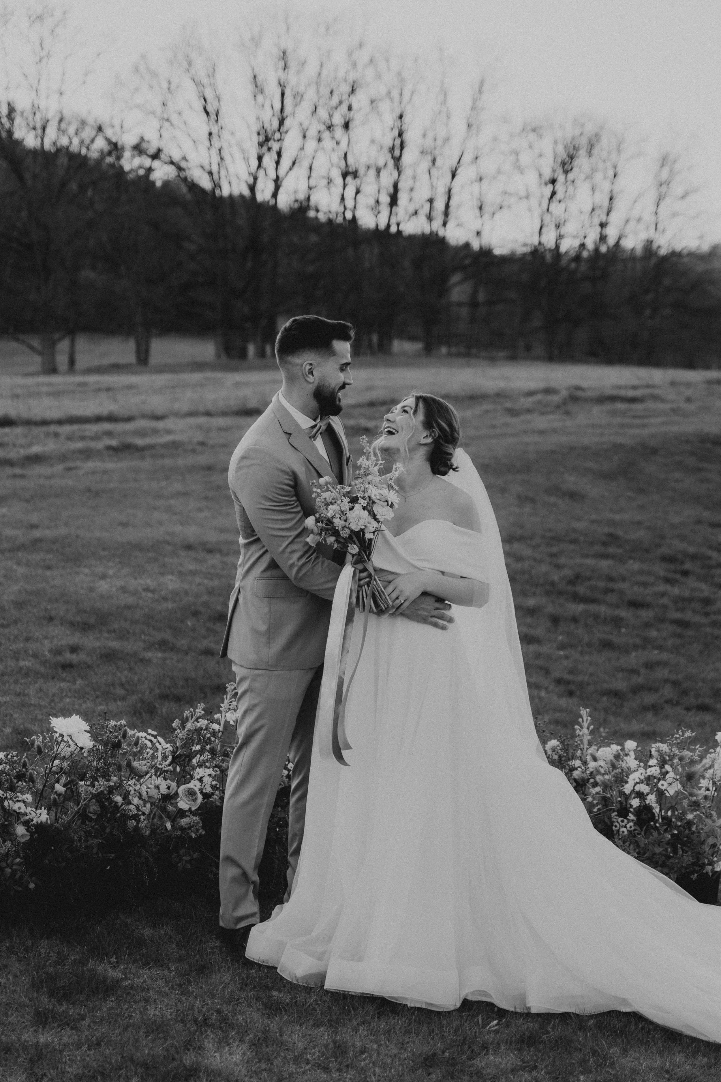A black-and-white photo of a bride and groom standing outdoors, smiling and looking at each other. The groom in a light suit holds a bouquet, and the bride in a flowing wedding dress is smiling with her hand on the groom's arm, surrounded by floral decorations and trees in the background.