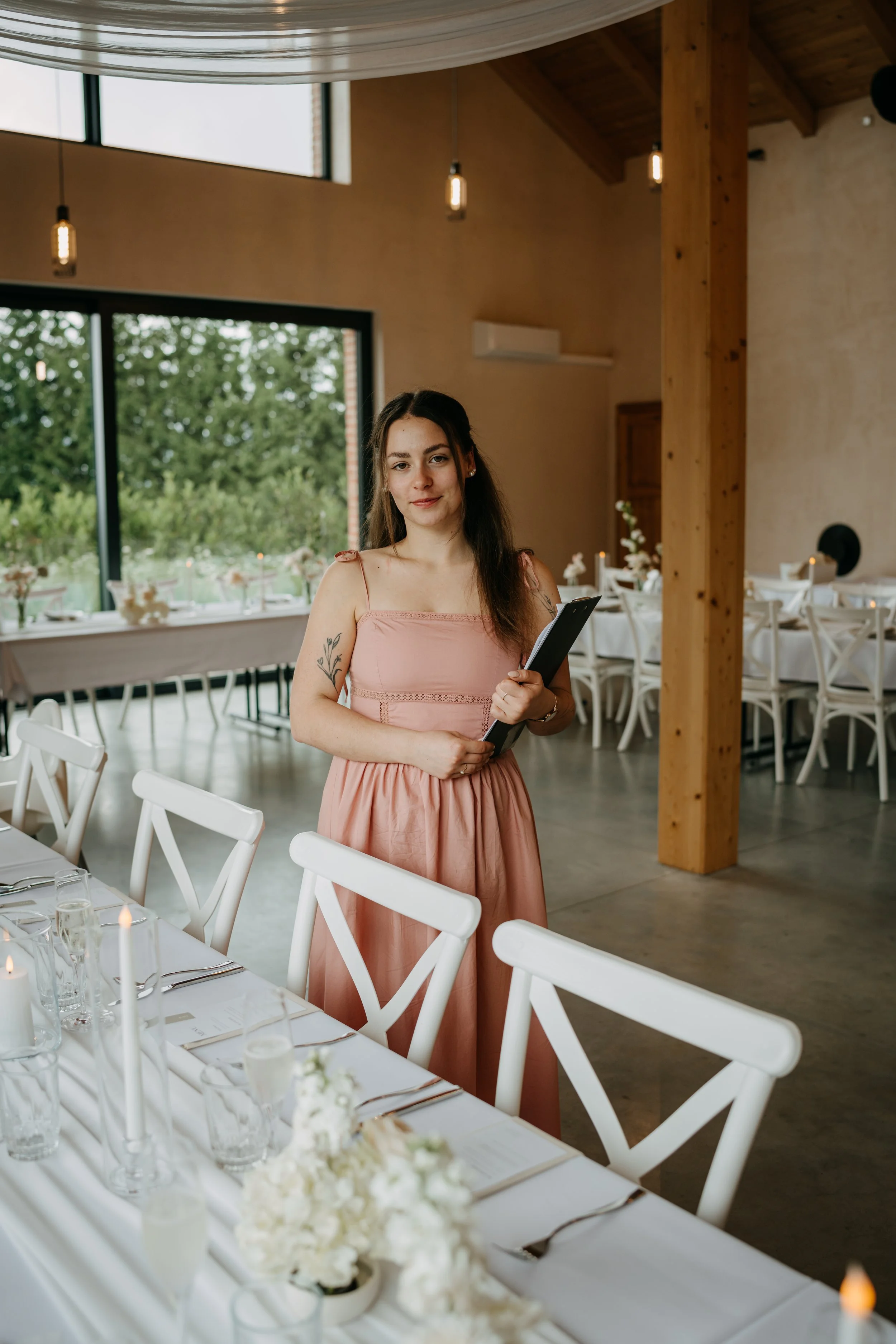 Young woman with long brown hair, wearing a sleeveless peach dress, holding a black folder, standing in a decorated event space with white chairs, dining table, and flower arrangements.