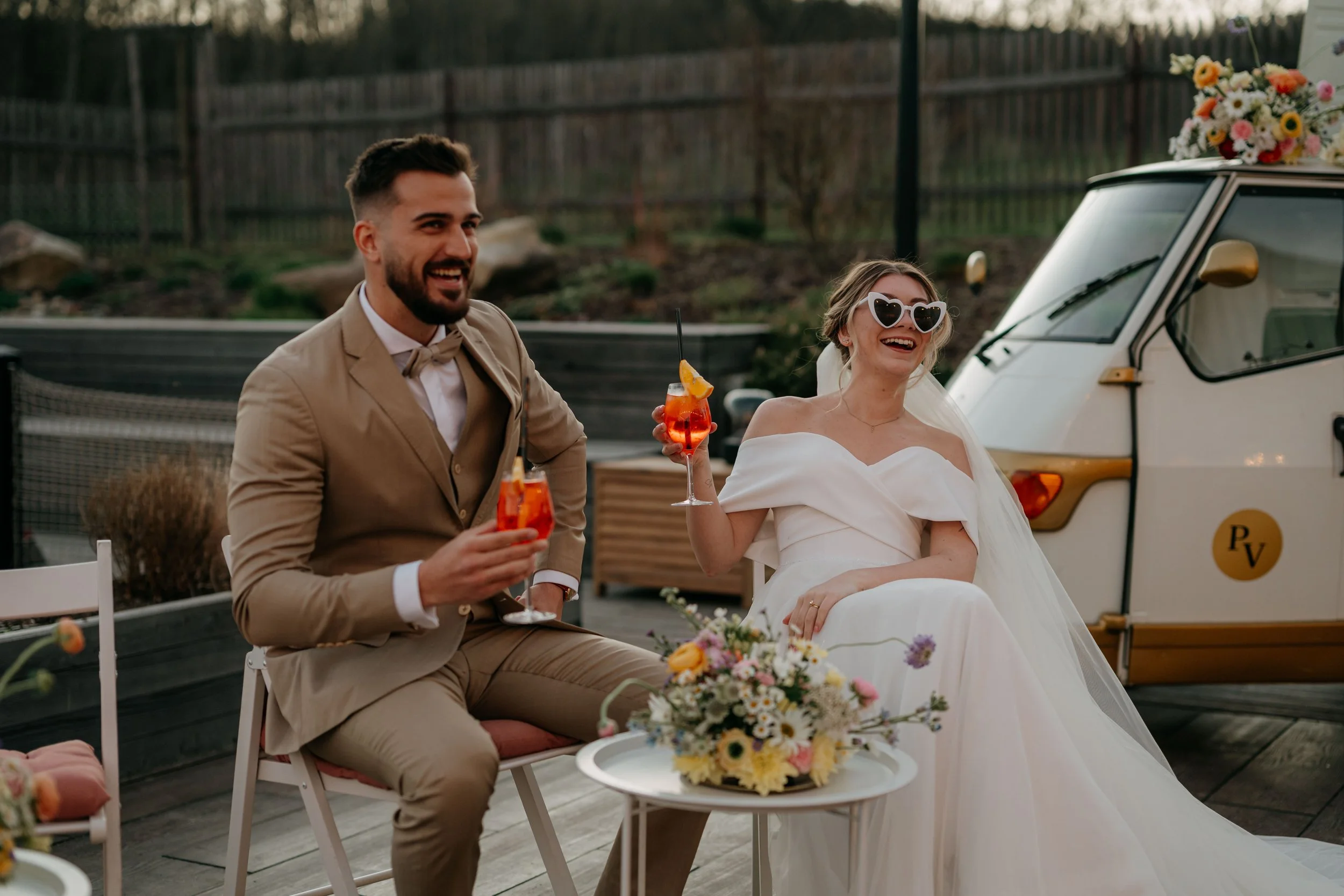 A bride and groom at a wedding reception, sitting outdoors with drinks, wearing formal attire, with a flower bouquet and a vintage vehicle in the background.