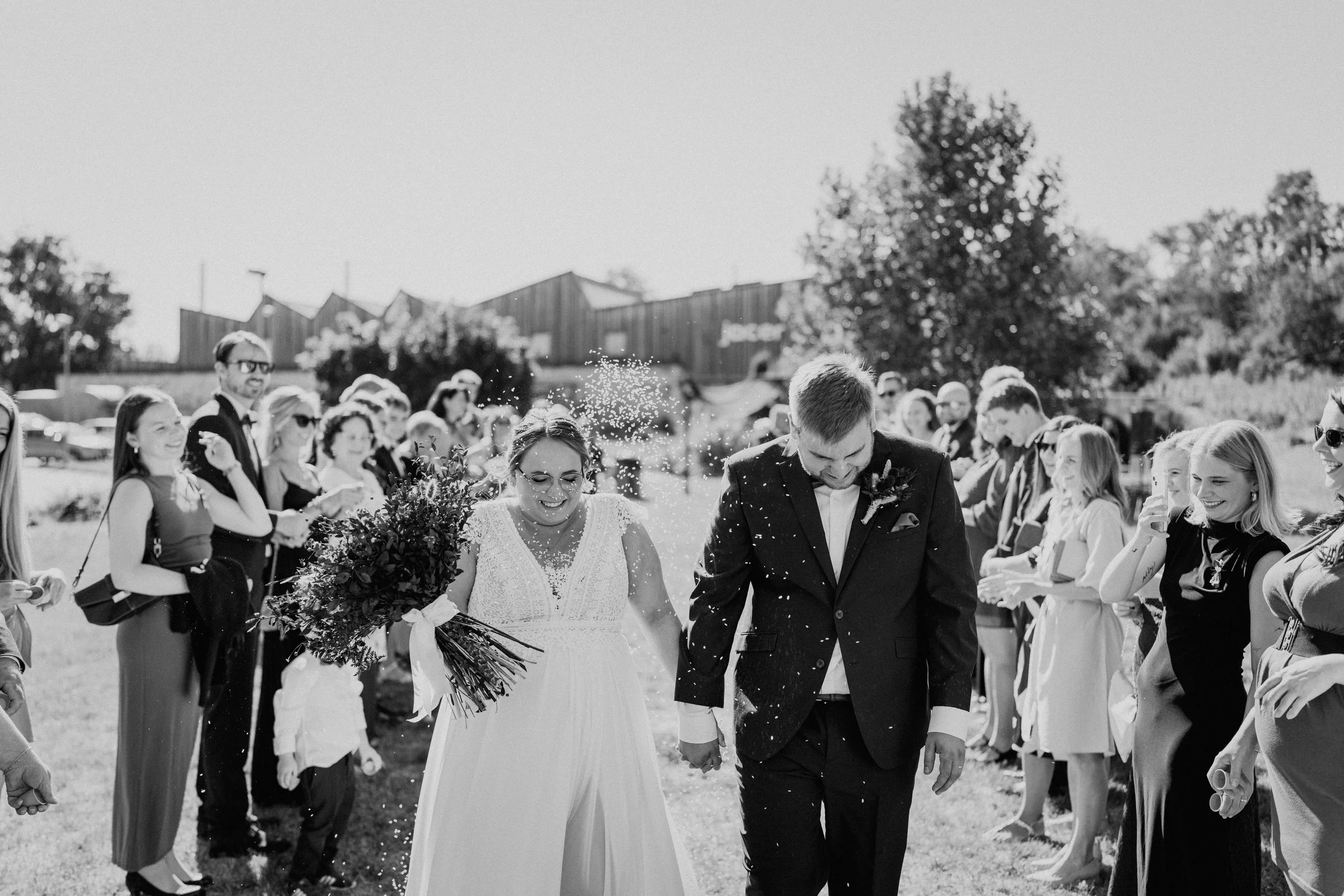Black and white photo of a wedding couple walking outdoors, surrounded by friends and family, with confetti falling around them.
