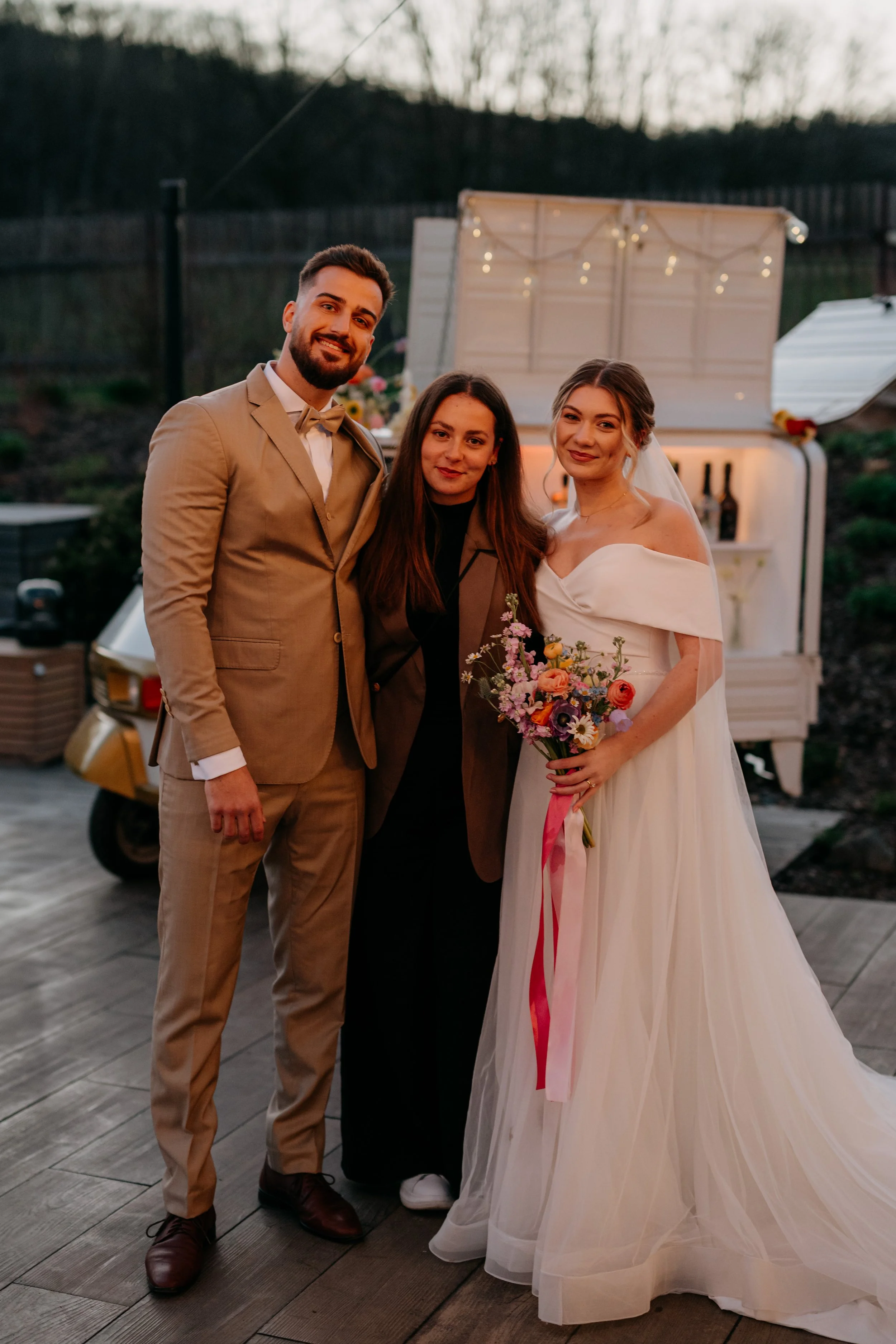 A wedding photo of three people outdoors during sunset. The bride is holding a flower bouquet, wearing a white wedding dress, and the groom is dressed in a tan suit. They are standing close together with a woman in black between them. There is a small white cart with string lights in the background.