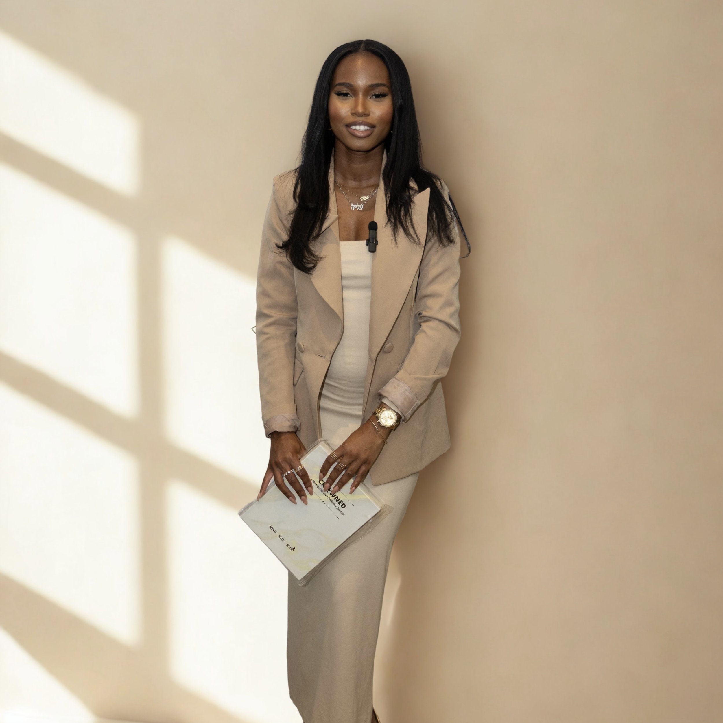 A woman in a beige suit holding a booklet, standing against a beige wall with sunlight casting shadows.