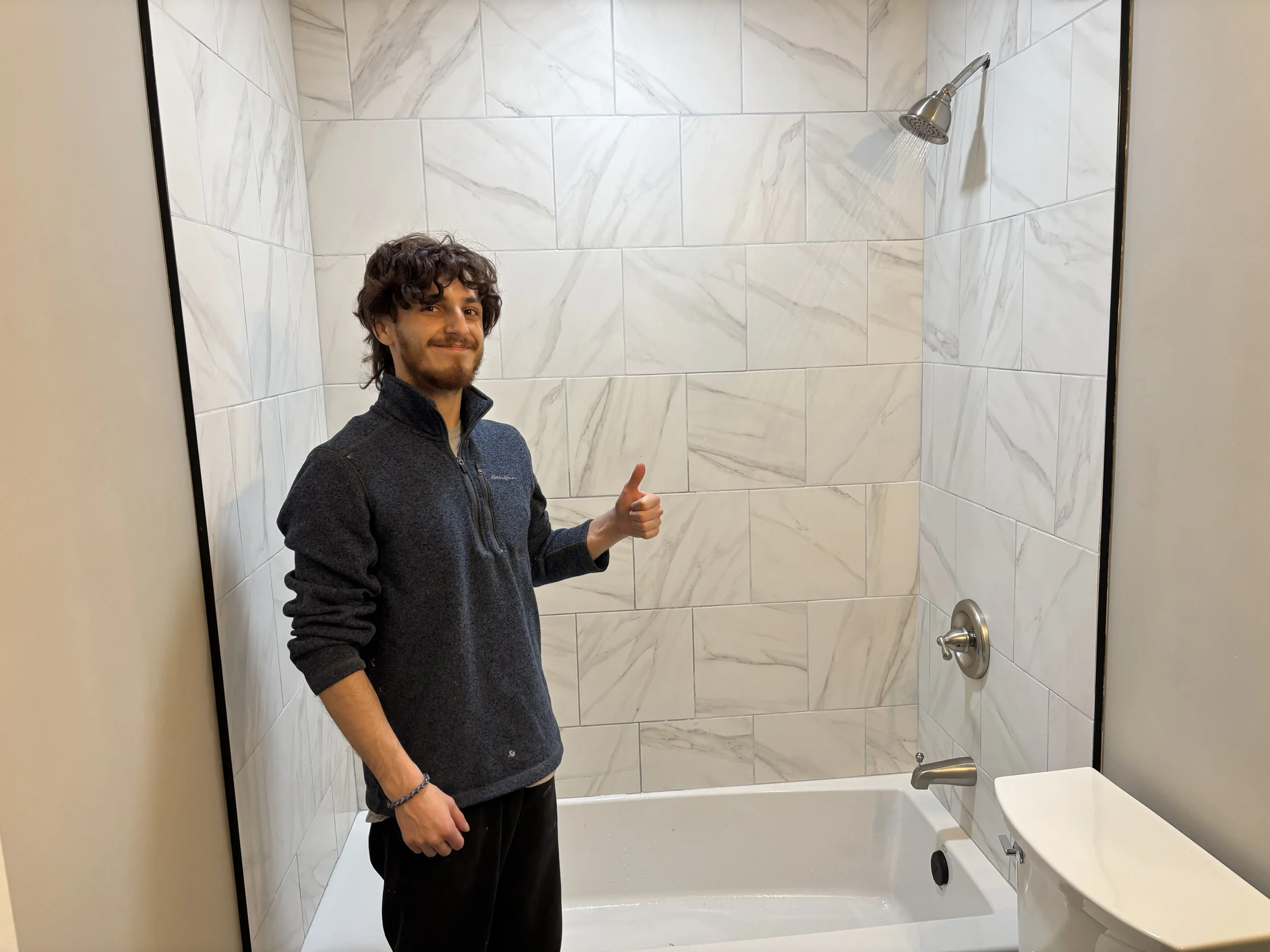 A young man with dark, curly hair and a beard, wearing a dark fleece jacket and black pants, is standing in a bathroom with a bathtub and shower. He is smiling and giving a thumbs-up gesture.