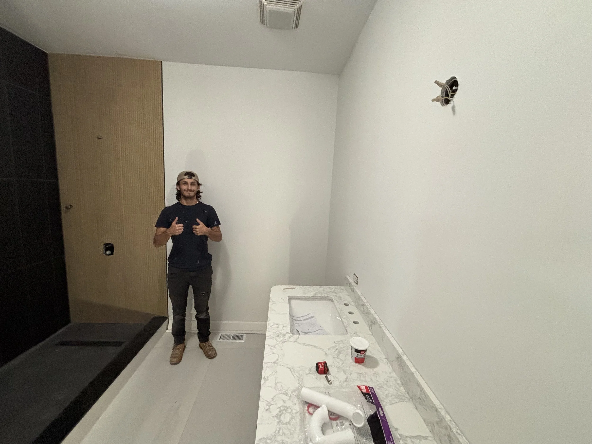 A man stands in a bathroom under renovation, giving a thumbs up. The bathroom has a marble countertop with an uninstalled sink, tools, and paint supplies on it. A partially installed light fixture is visible on the wall opposite the man. The walls are white, and there's a wooden panel next to a black feature wall.