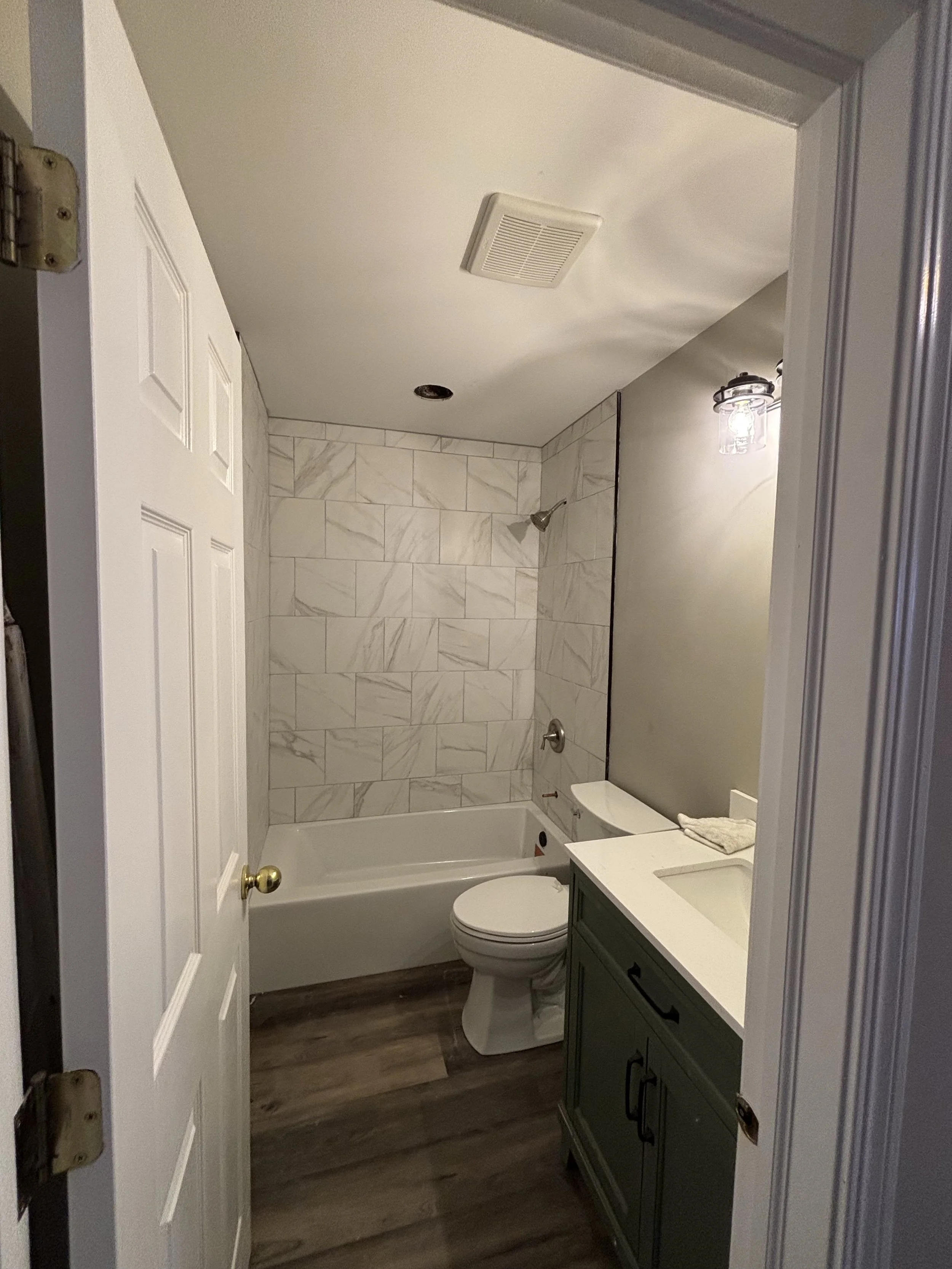 A small bathroom with a bathtub and shower, a toilet, and a green vanity with a white sink. The walls have gray paint and tile, and there is a light fixture above the sink.