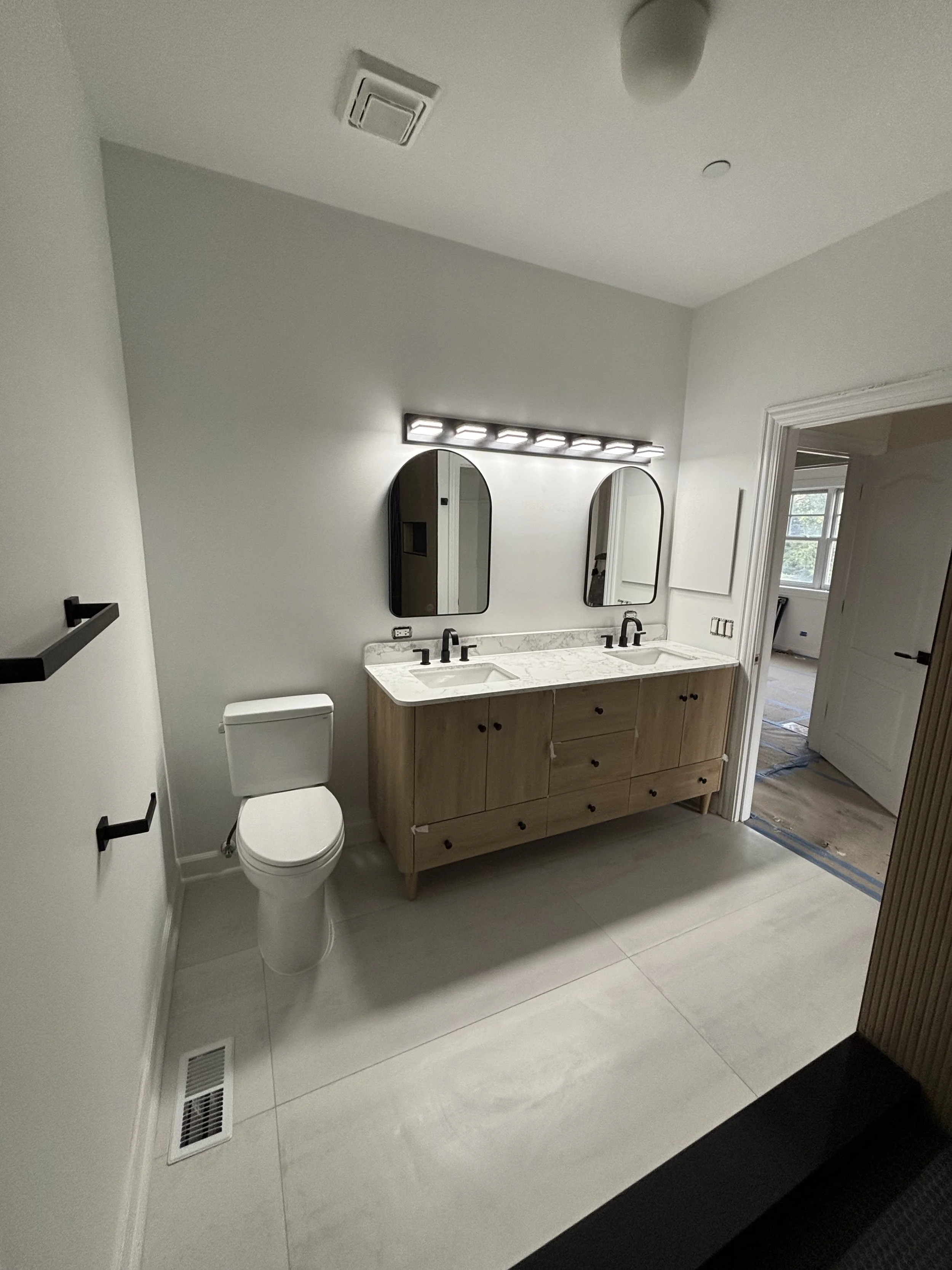 Modern bathroom with dual sinks, black fixtures, two oval mirrors, white walls, light-colored flooring, and a toilet.