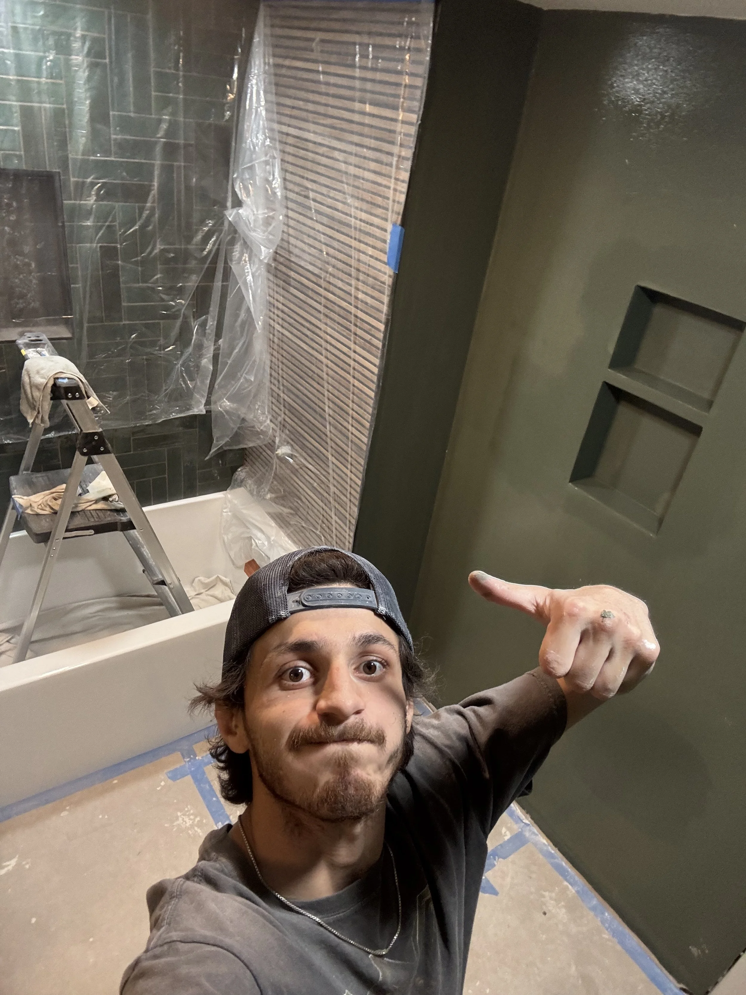 A man with a baseball cap taking a selfie in a bathroom under renovation. The bathroom has a partially tiled wall, a bathtub, a ladder, and plastic sheeting on the wall.