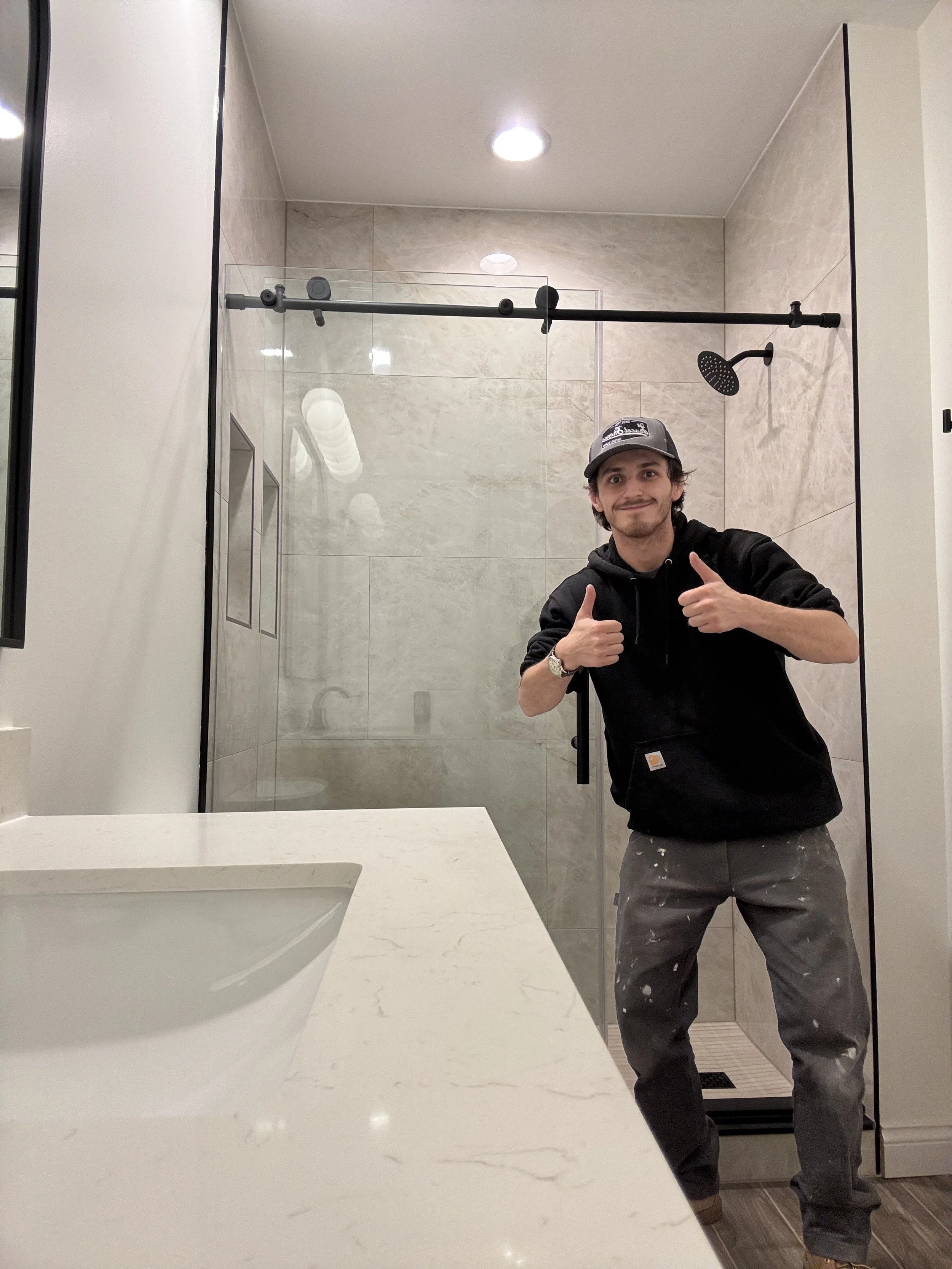 A man in a black hoodie and gray pants standing in a modern bathroom shower area, giving a thumbs-up gesture.