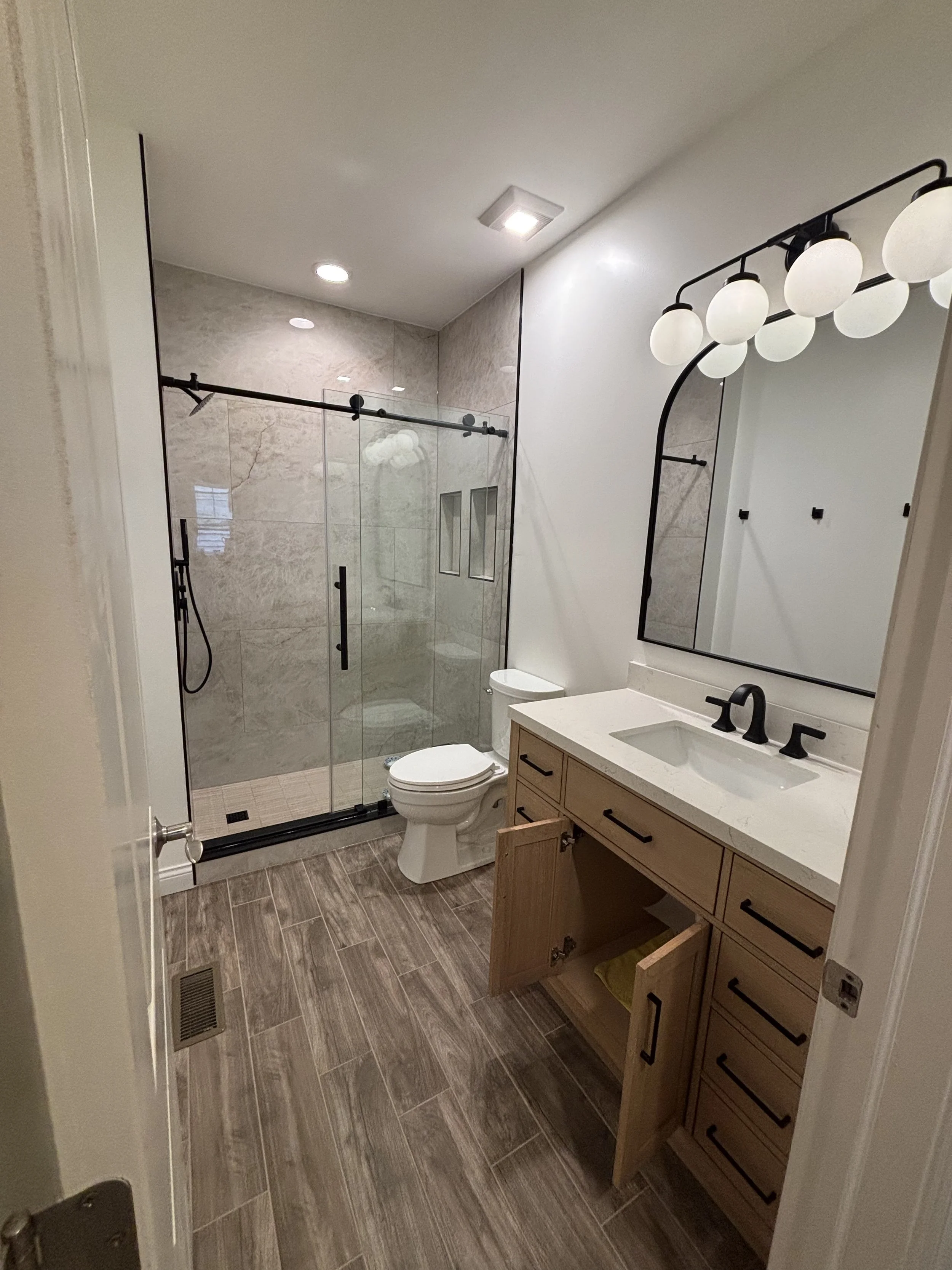 Modern bathroom with a walk-in shower, toilet, wooden vanity with a sink, and a large mirror with round globe lights. The floor is wood-look tile, and the shower has glass doors and beige tiles.