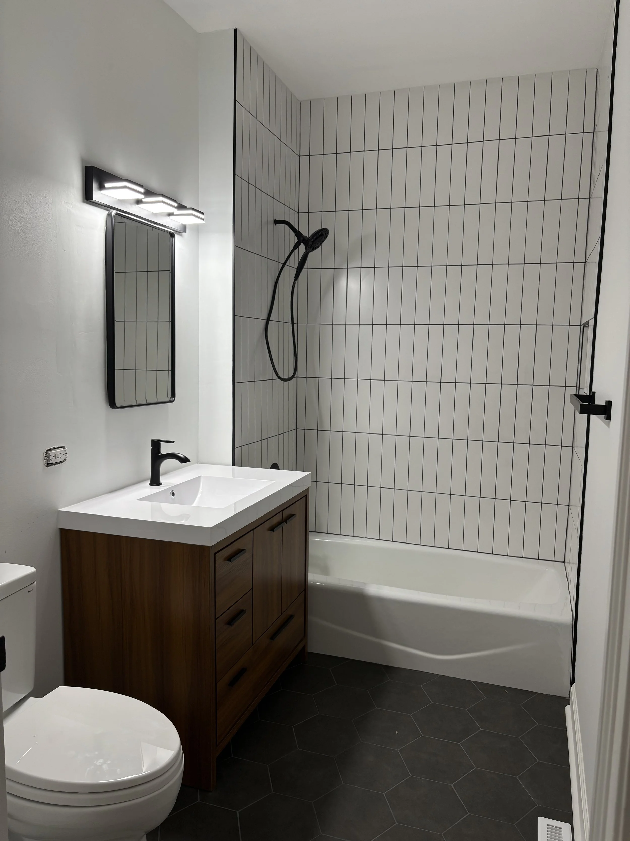Modern bathroom with a white bathtub and shower area, black showerhead, white tiled wall with black grout lines, wooden vanity with a white sink, black faucet, and a mirror with a black frame. The floor has dark hexagon tiles.