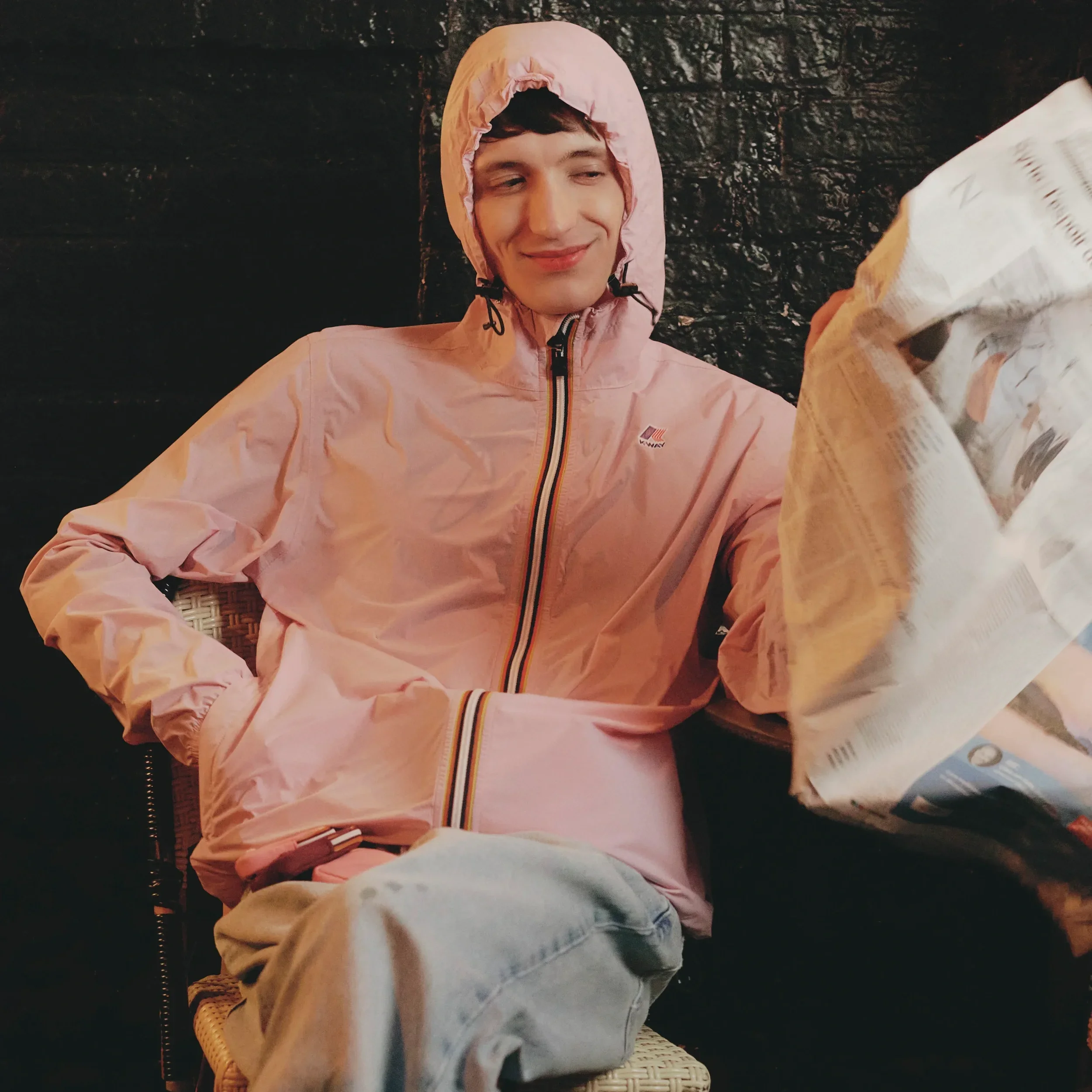 Uomo indossa giacca a vento K-Way rosa con cappuccio e iconica zip a righe, jeans in denim chiaro dal taglio morbido e sneakers bianche.