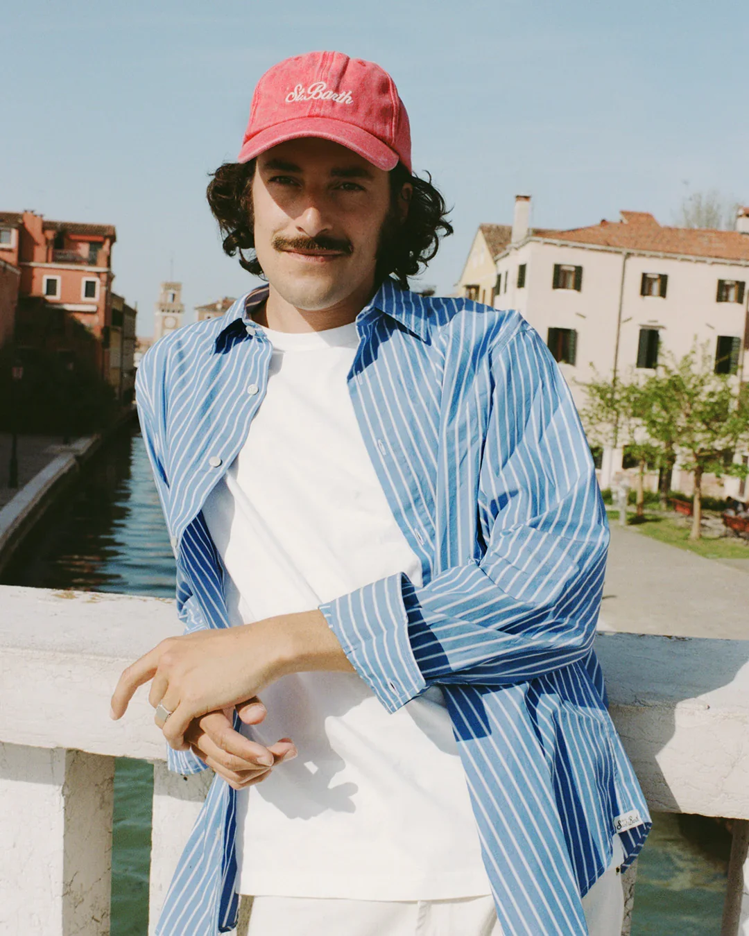 Uomo con baffi e capelli mossi indossa una camicia a righe verticali azzurre e bianche aperta sopra una t-shirt bianca, pantaloni chiari e un cappellino da baseball rosso Saint Barth con ricamo bianco
