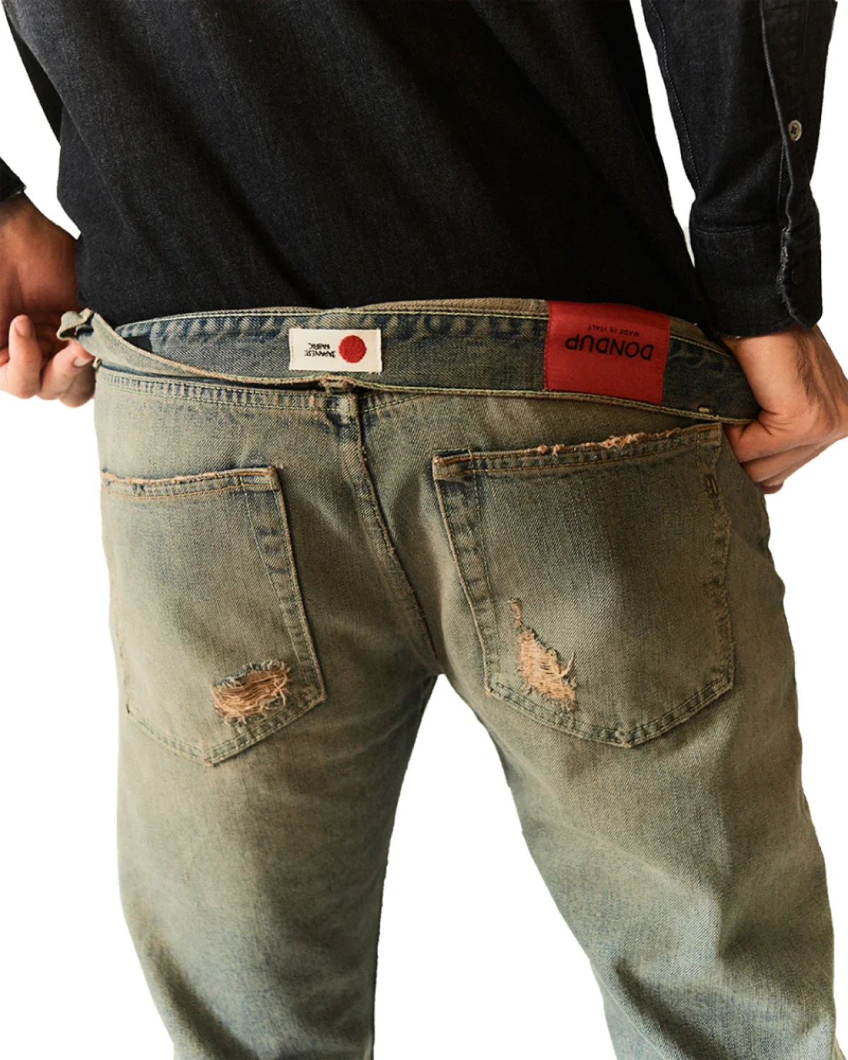 Dettaglio posteriore di jeans Dondup uomo in denim chiaro slavato con effetto sporco e strappi, etichetta rossa Dondup e camicia di jeans scura.