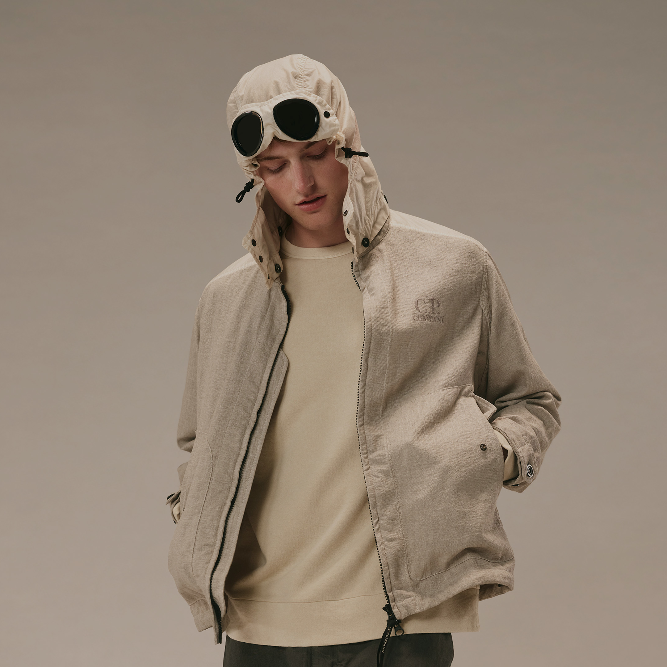 Uomo indossa giacca leggera beige C.P. Company con cappuccio e lenti integrate, t-shirt girocollo color panna e pantaloni scuri.