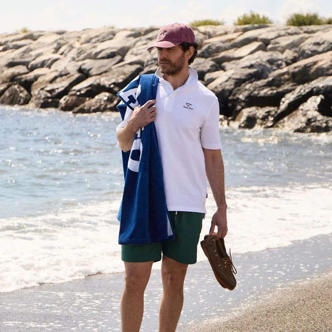 Uomo indossa polo bianca Sebago, pantaloncini mare verdi, cappellino con visiera bordeaux e tiene in mano un telo mare blu e classiche scarpe da barca in pelle marrone.