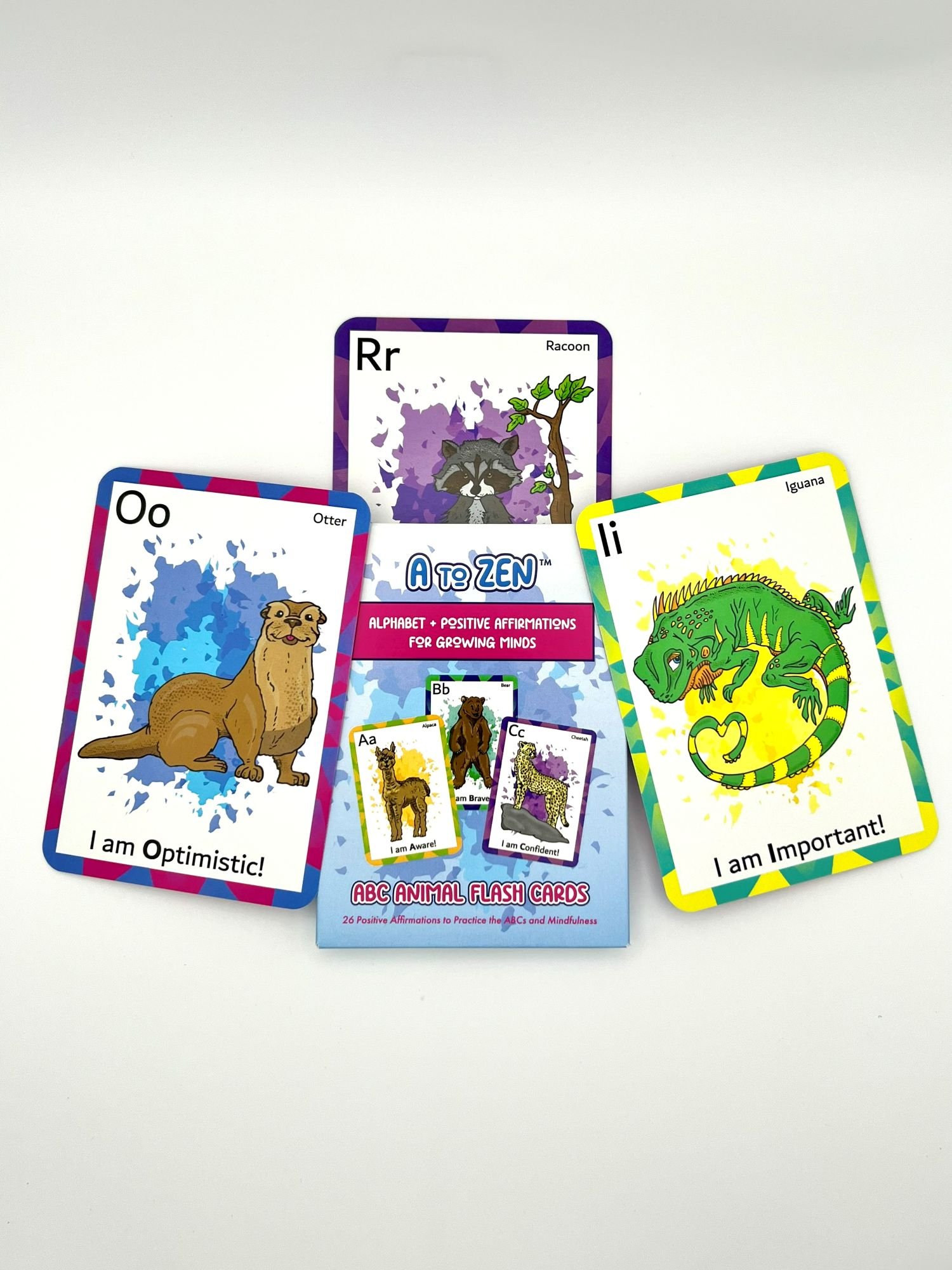 Set of three colorful flash cards with animal illustrations and positive affirmations, including an otter with 'I am Optimistic!', a raccoon with 'I am Aware!', and a green iguana with 'I am Important!', beneath a box of alphabet flash cards.