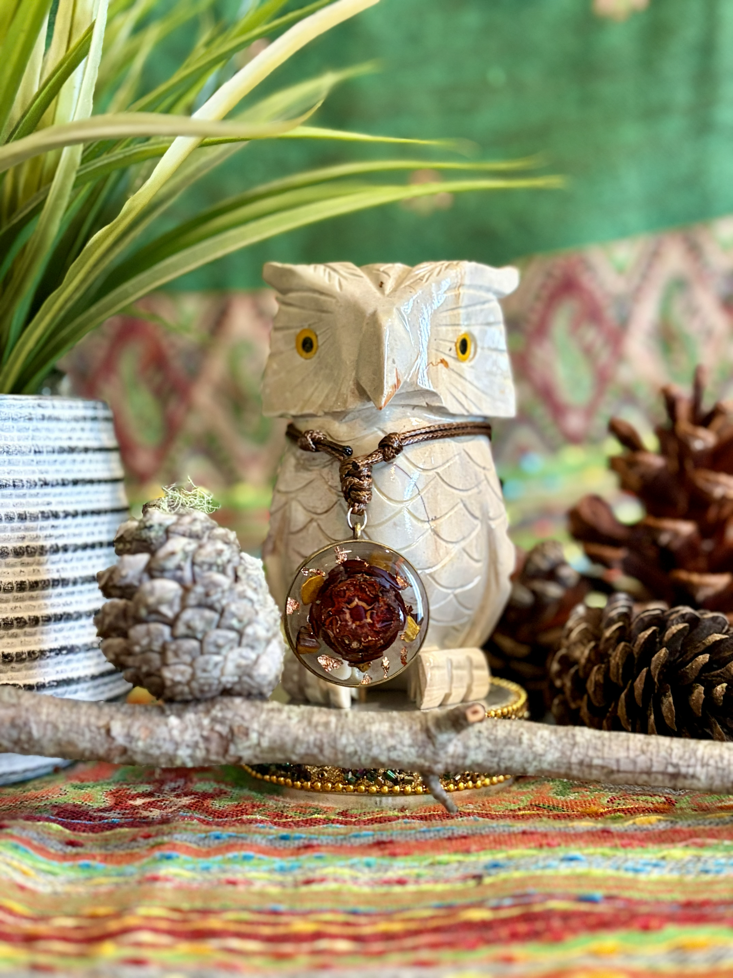 Decorative owl ceramic figurine with a necklace hanging around its neck, surrounded by pinecones, pinecone, a branch, and a potted plant in a striped pot, on a colorful woven cloth.