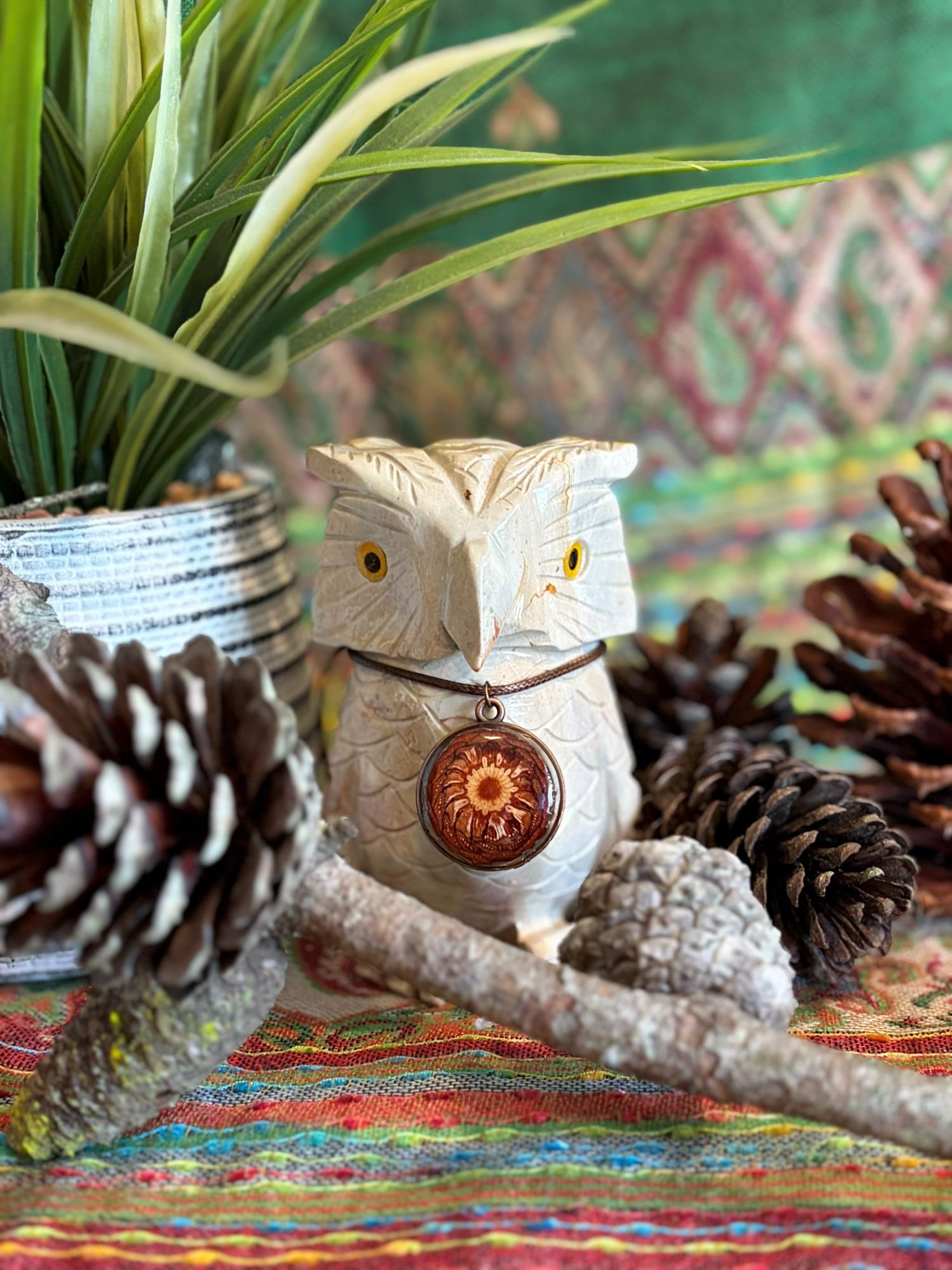 Decorative owl sculpture with yellow eyes and a circular pendant around its neck, surrounded by pinecones, a plant, and colorful fabric background.