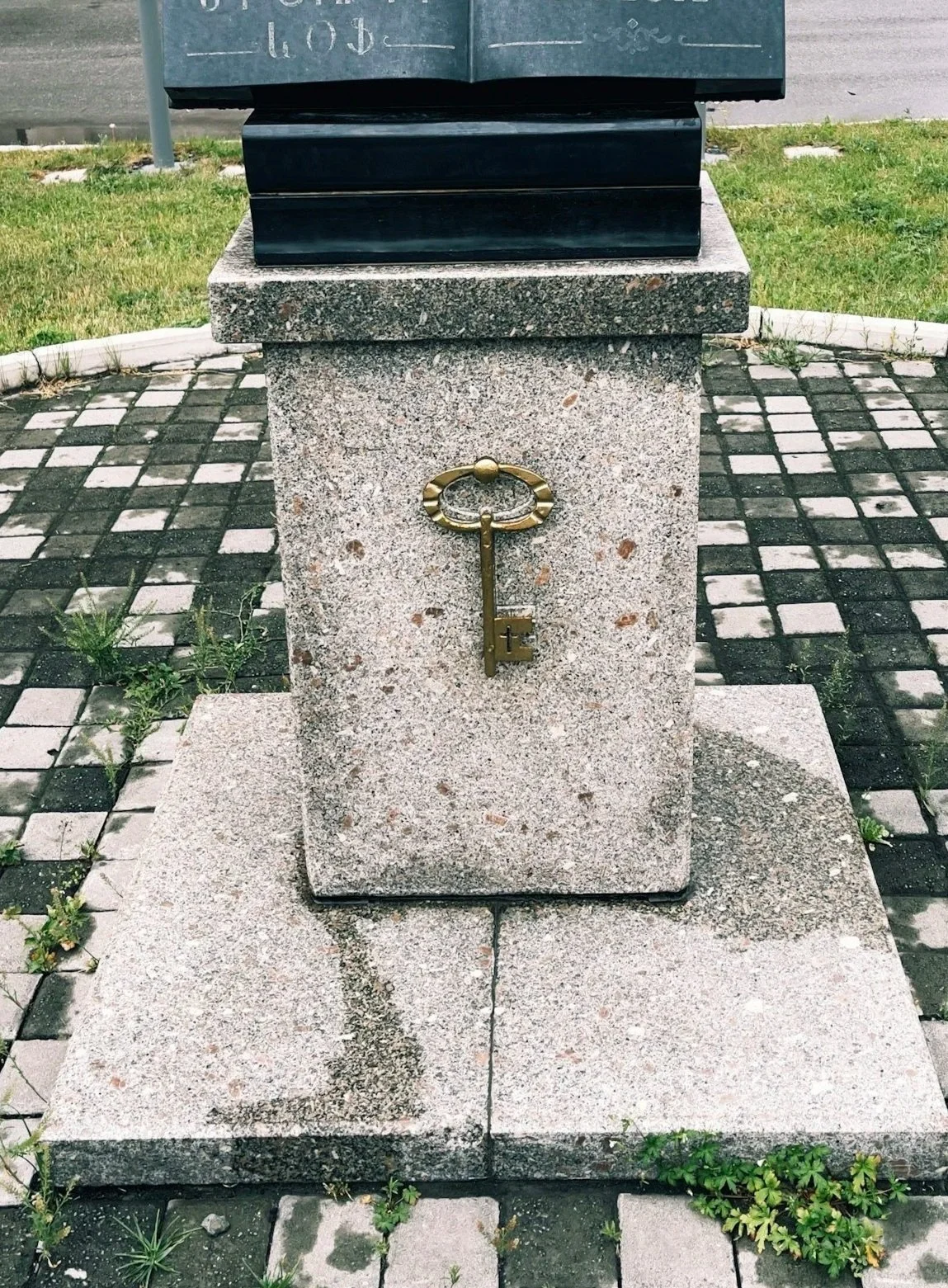 A memorial stone with a brass key attached to it, placed on a concrete base on a cobblestone pavement with grass around.