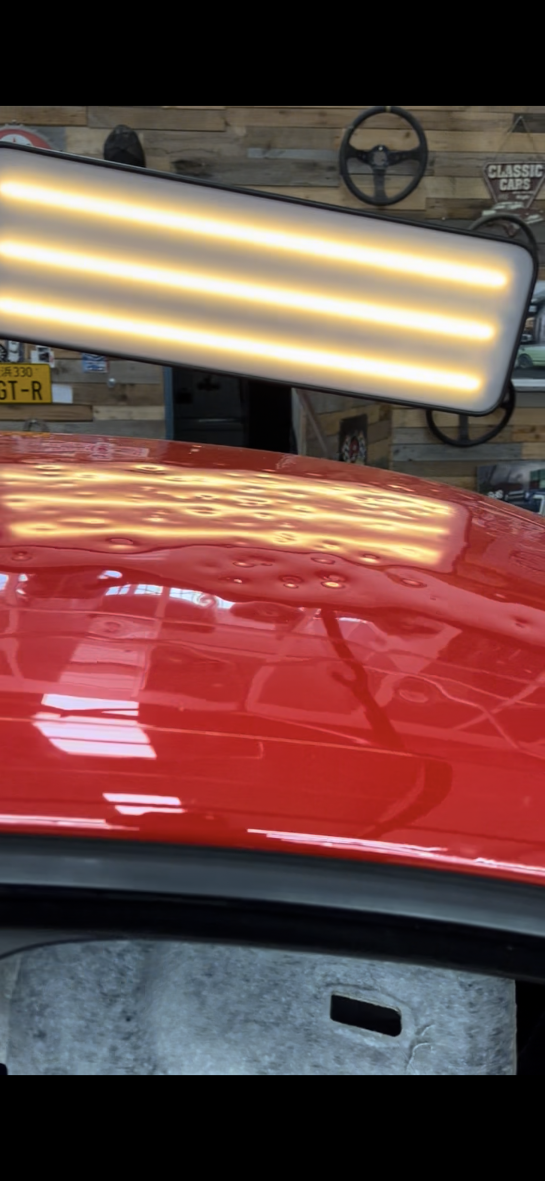 A red car with a shiny surface reflecting a large rectangular LED light fixture, a steering wheel, and vintage license plates in the background.